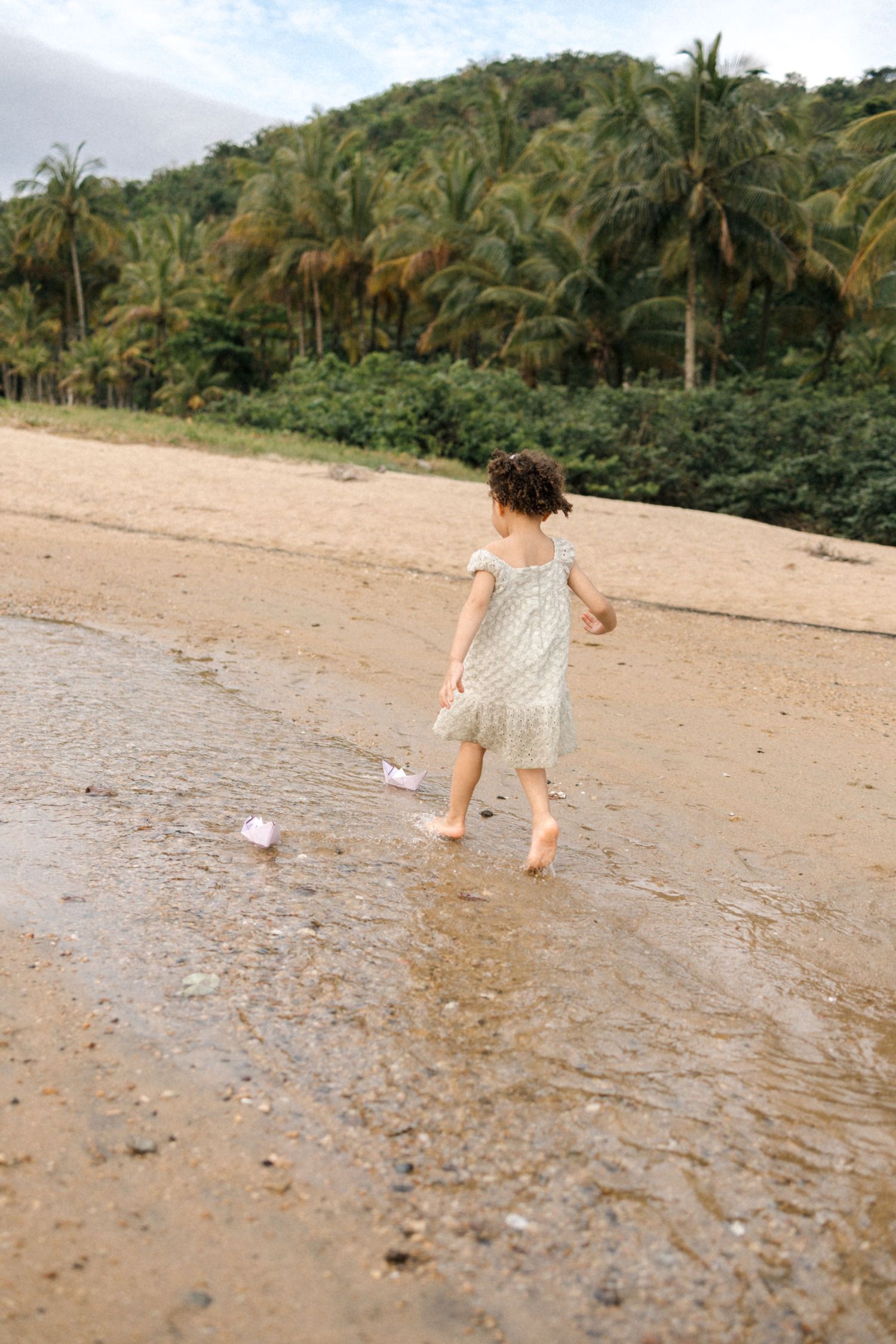 Child running along a tropical beach with lush palm trees and mountains in the background.