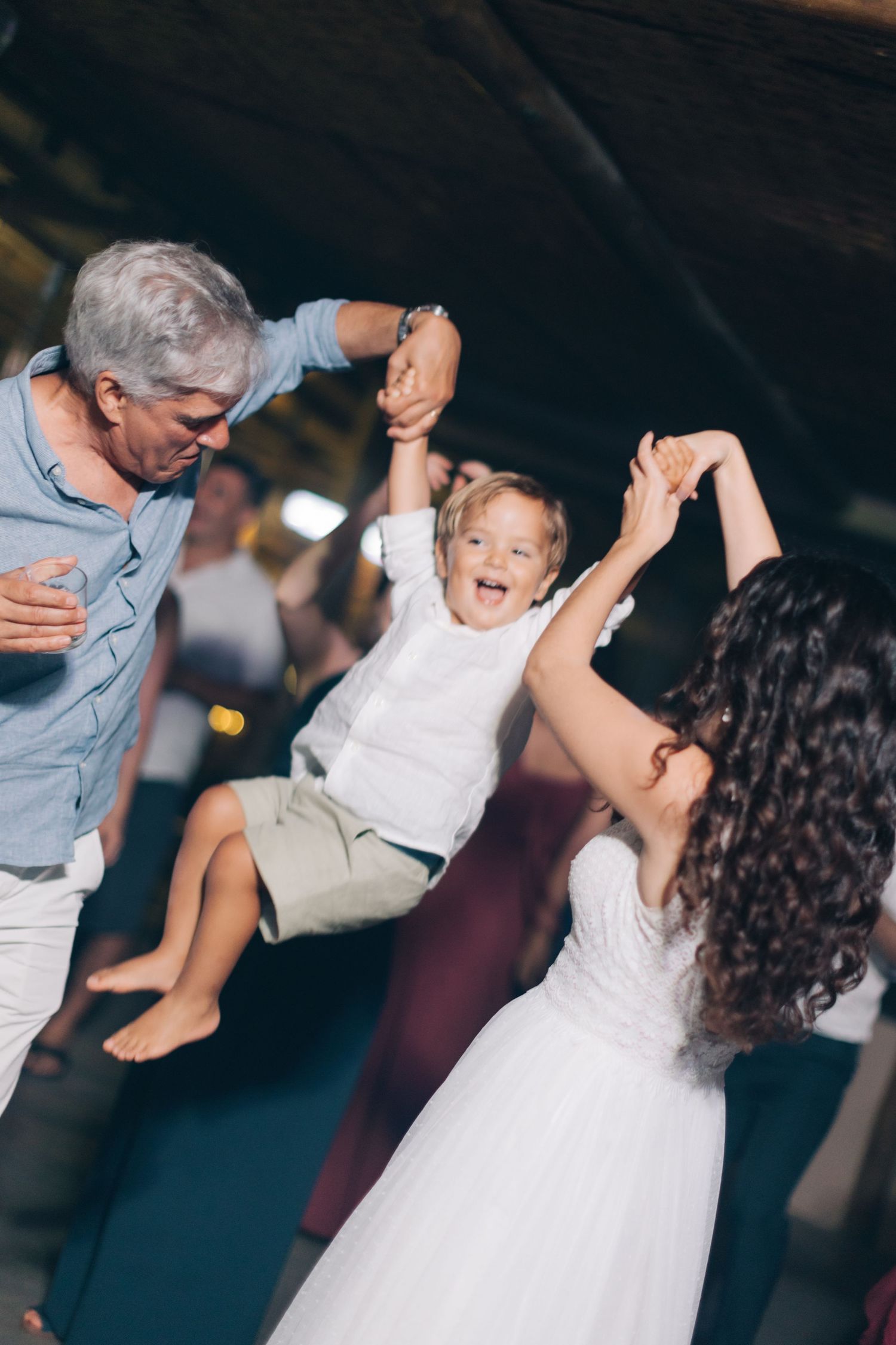 Adults swing a small child between them while dancing at a wedding celebration.