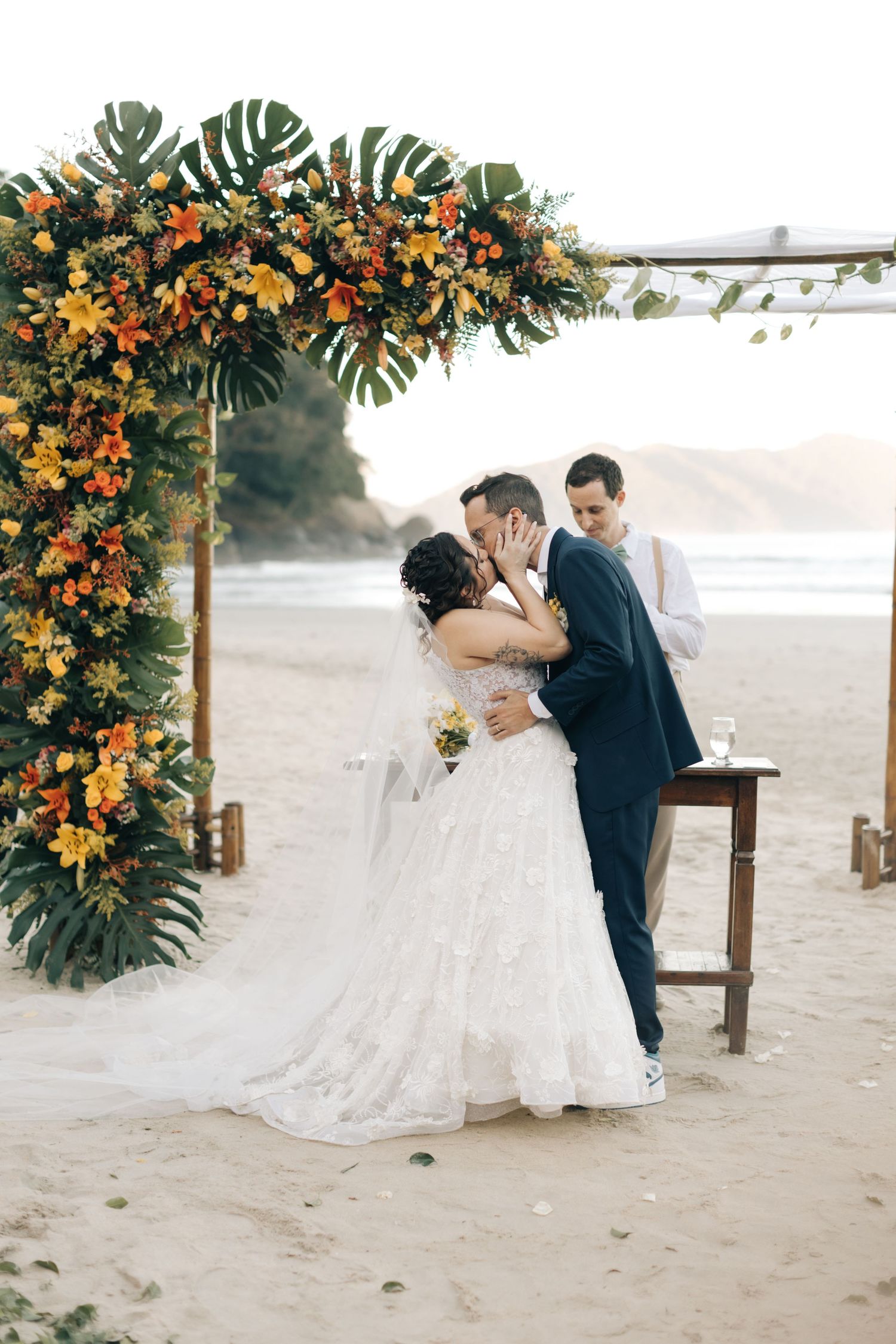 Couple kisses beneath tropical floral arch during beach wedding ceremony.