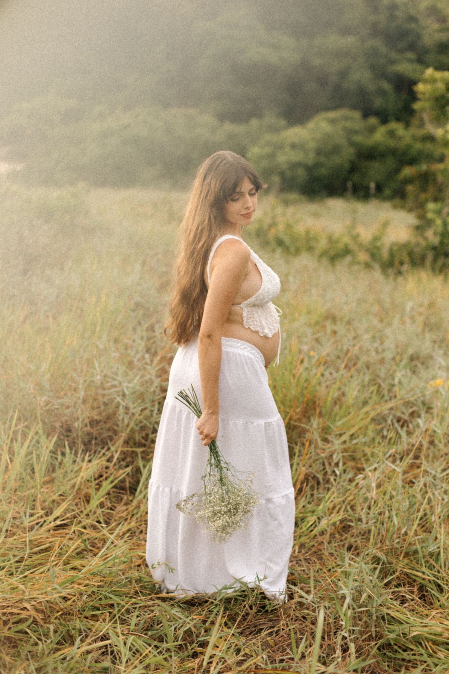 Ethereal outdoor maternity photo of woman in white dress holding wildflowers in a meadow.