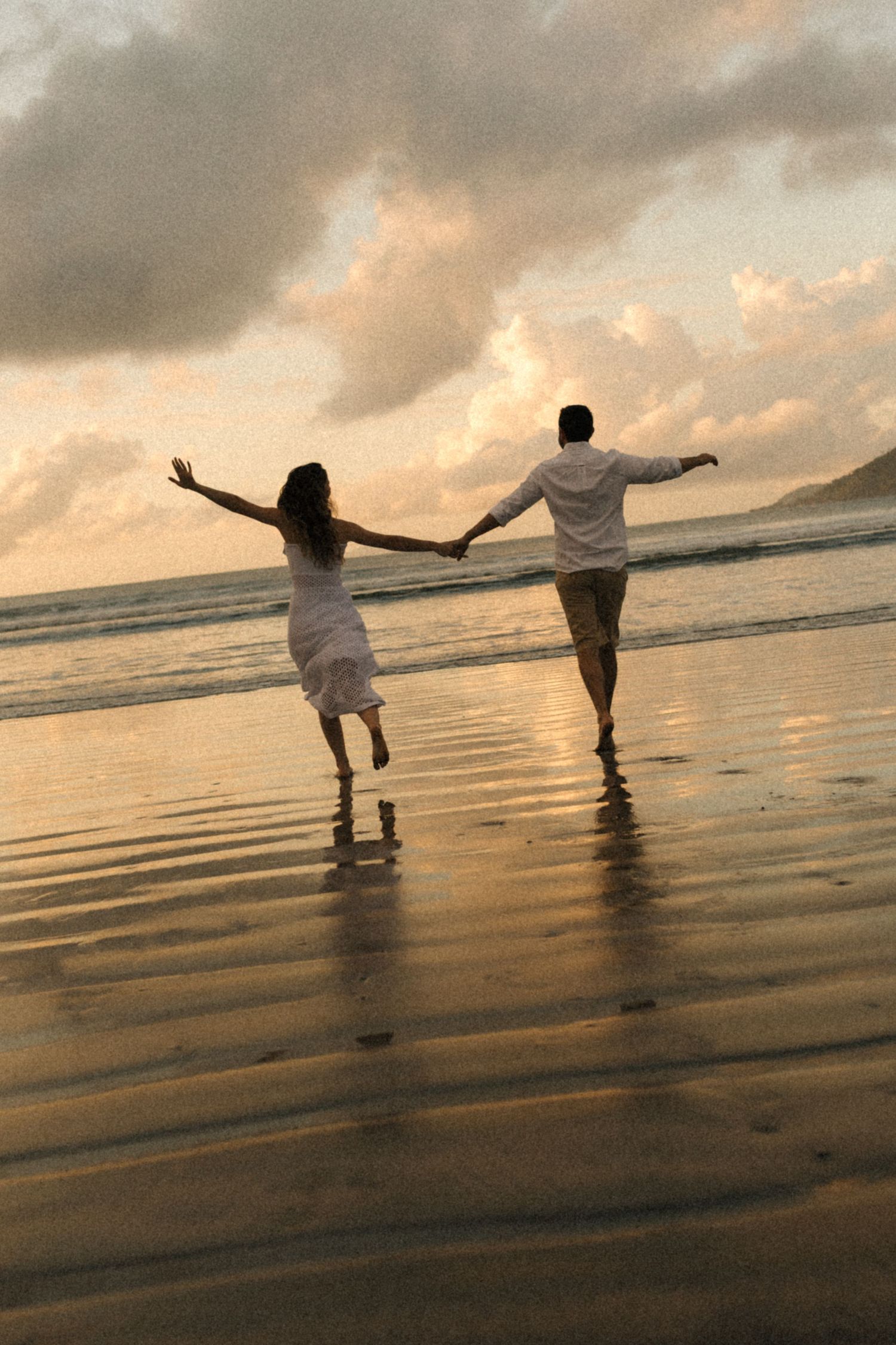 Two people spread their arms in joy while walking through beach waters at sunset.