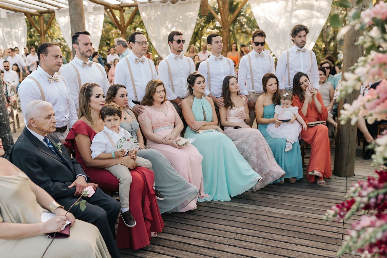 Wedding guests seated in rows on wooden deck wearing pastel and formal attire.