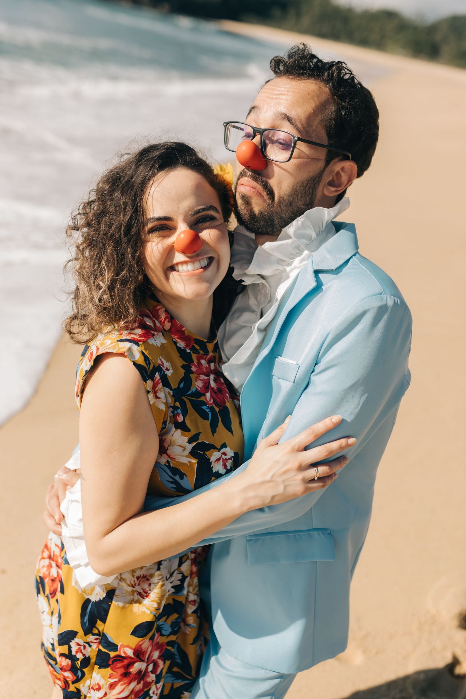 A couple wearing red clown noses embrace on a sunny beach with ocean waves in the background.