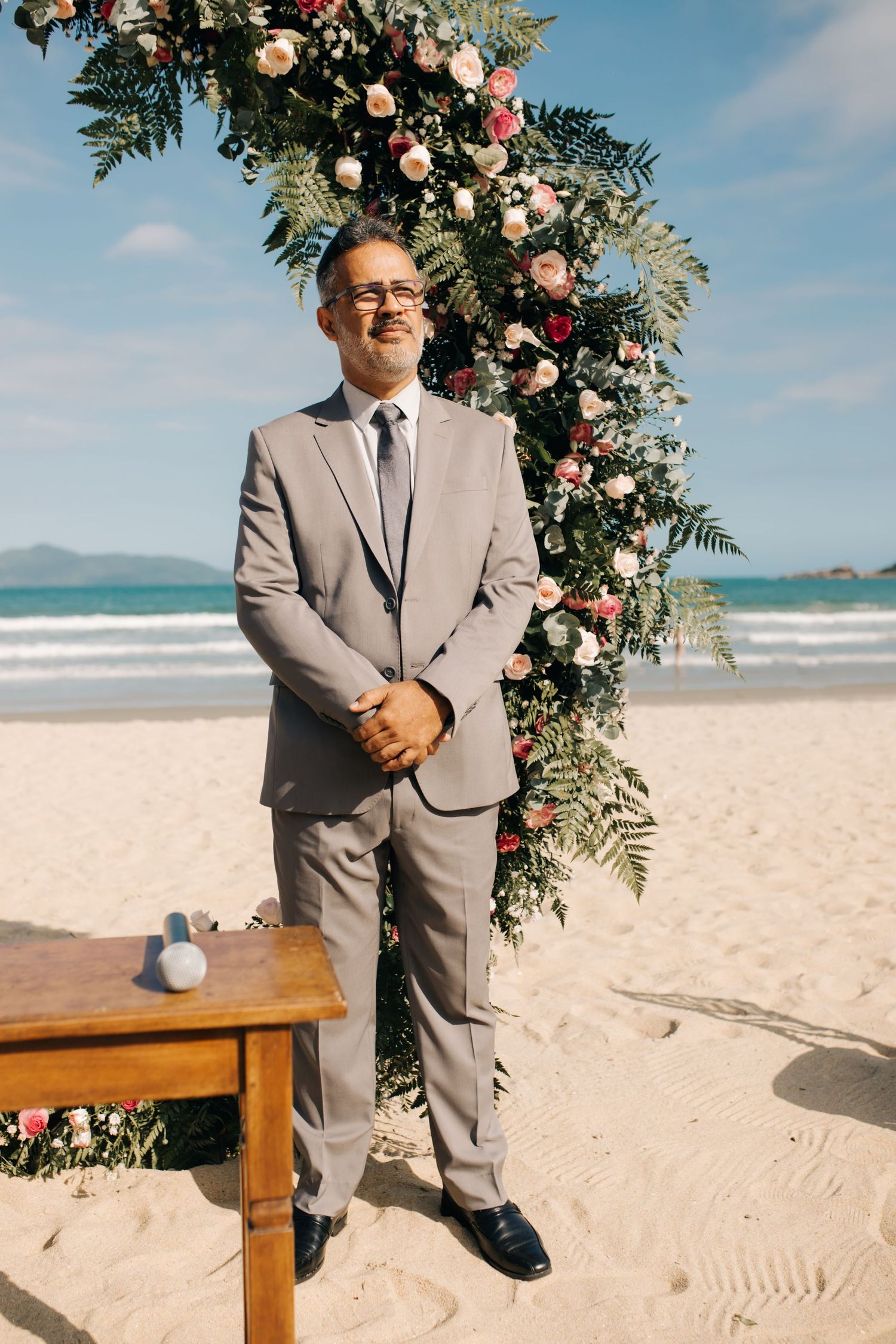 A person in a gray suit stands beside a floral arch with pink roses on a sunny beach.