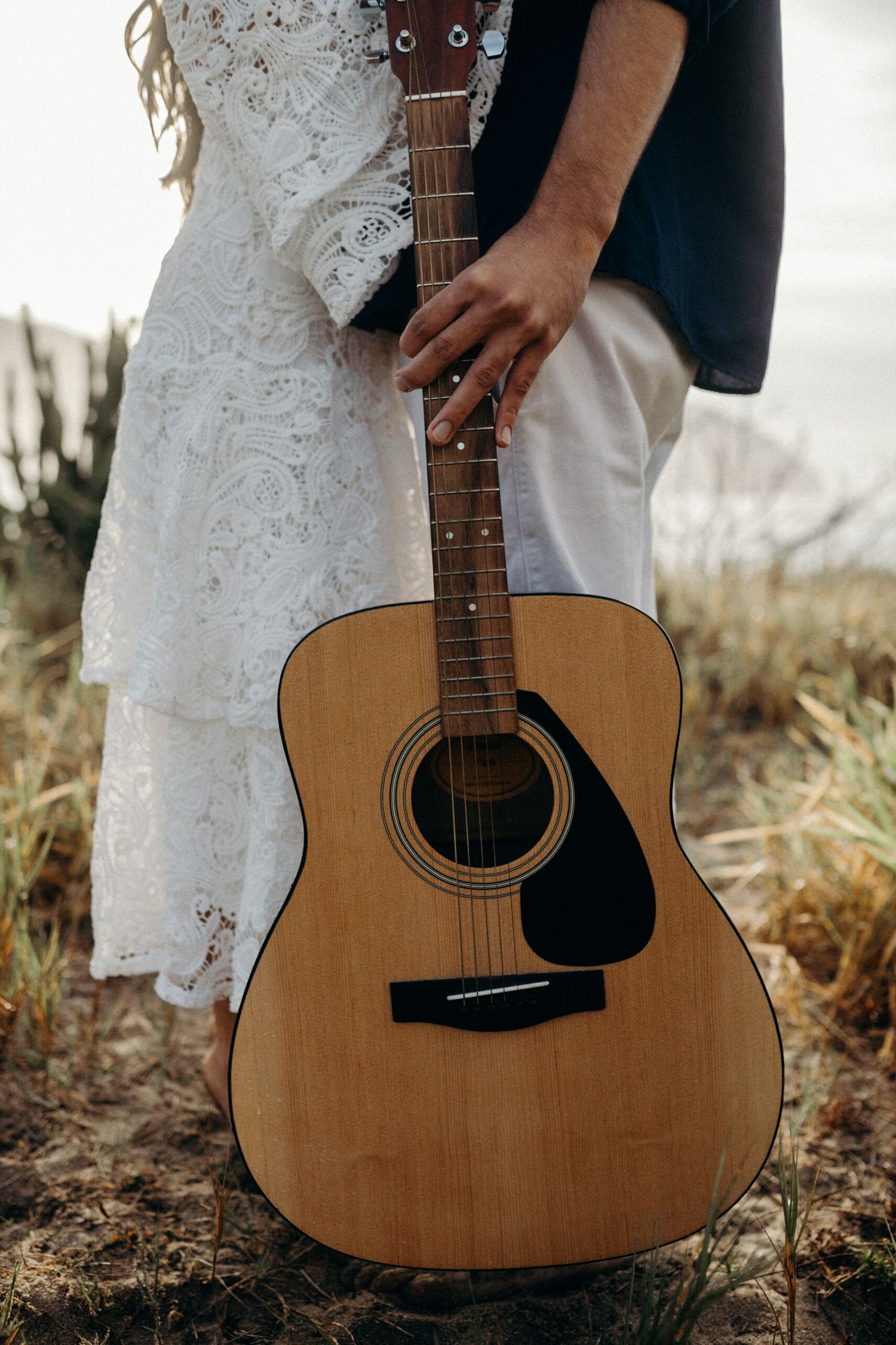 An acoustic guitar stands prominently against a white lace dress and dark clothing in a rustic outdoor setting.