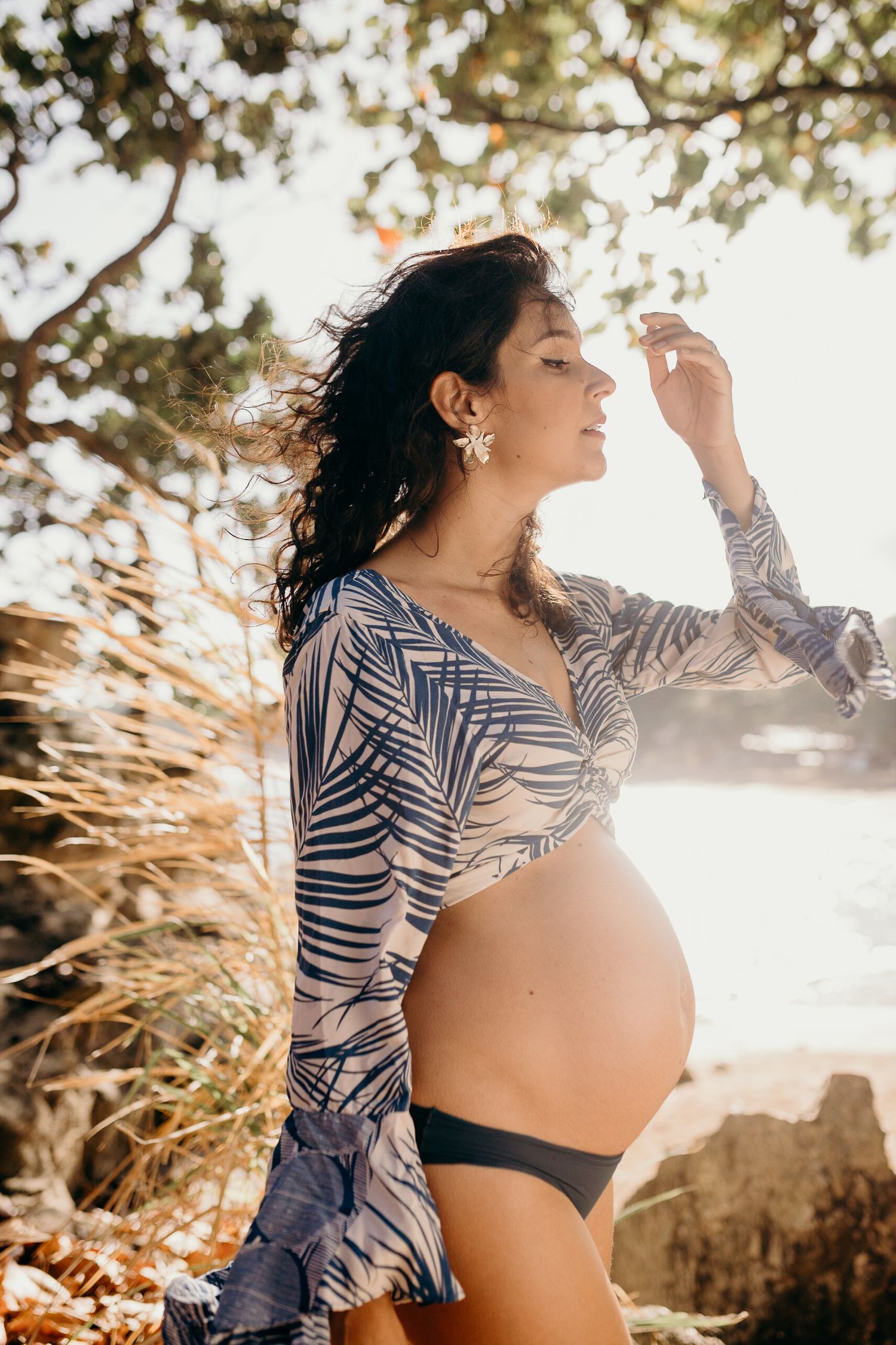 A backlit maternity portrait at sunset with palm fronds and rocky coastline.