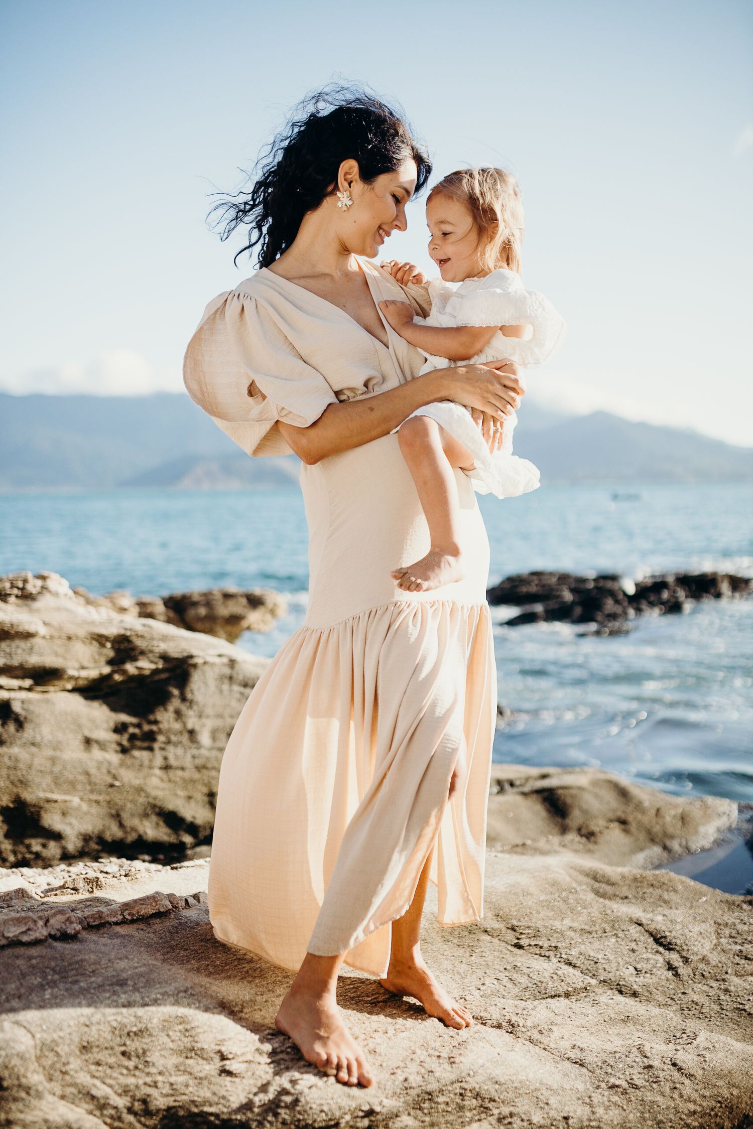Parent and child sharing tender moment on rocky coastline while wearing light colored flowing dress.