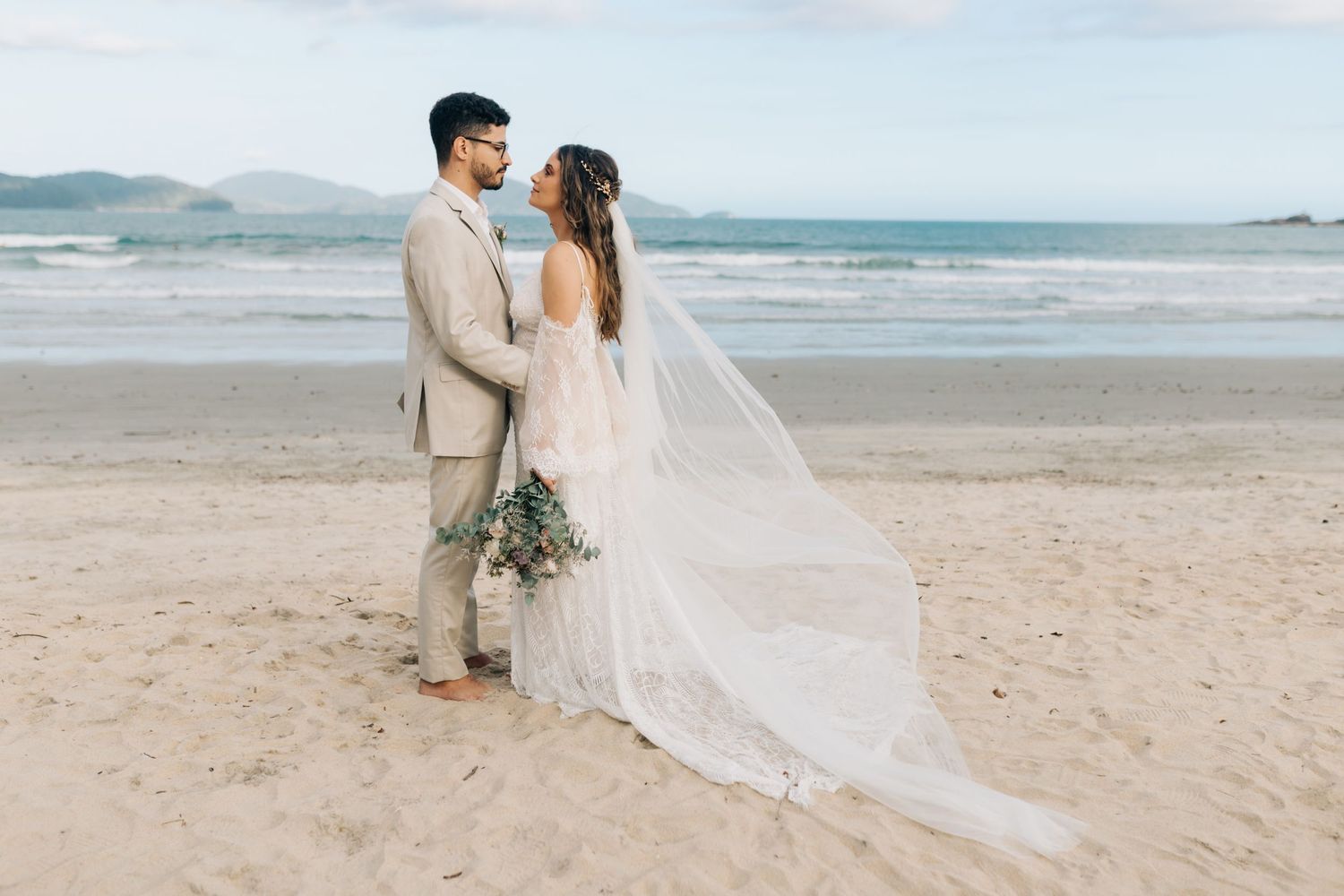 Romantic beach wedding portrait with flowing white veil against coastal landscape.