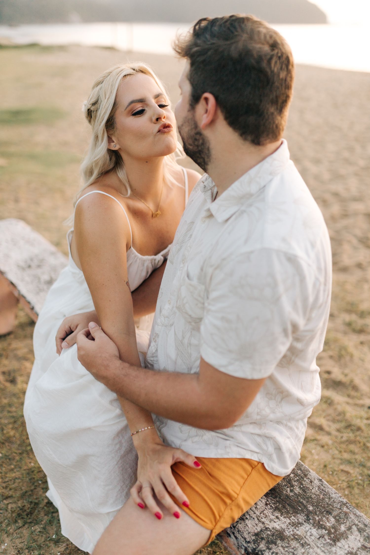 An intimate moment between a couple sitting on driftwood at a scenic beach during sunset.