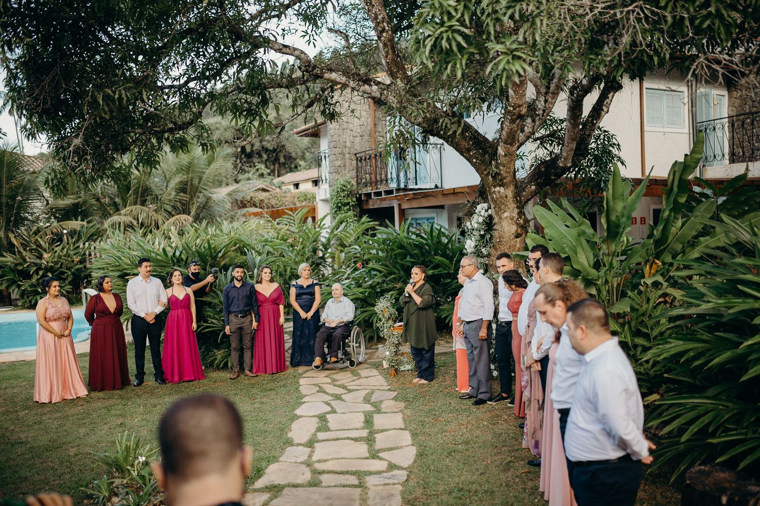 Wedding guests line stone pathway surrounded by tropical garden landscaping and pool.