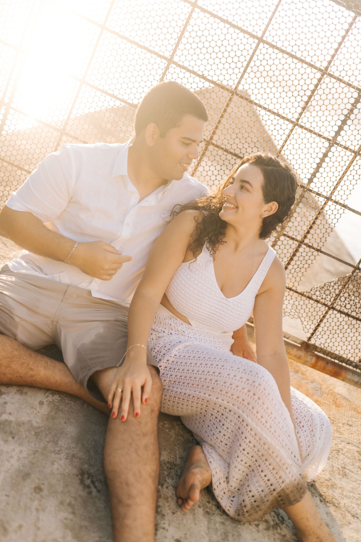 A couple sharing laughter while sitting together against a sun-lit fence backdrop.
