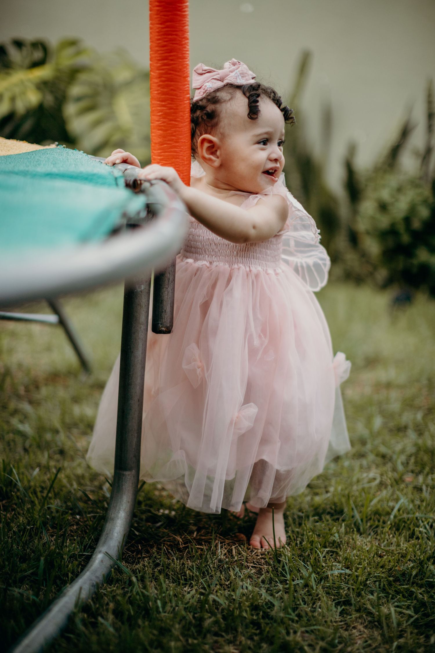 Young child in pink tulle dress smiling while standing next to furniture outdoors.