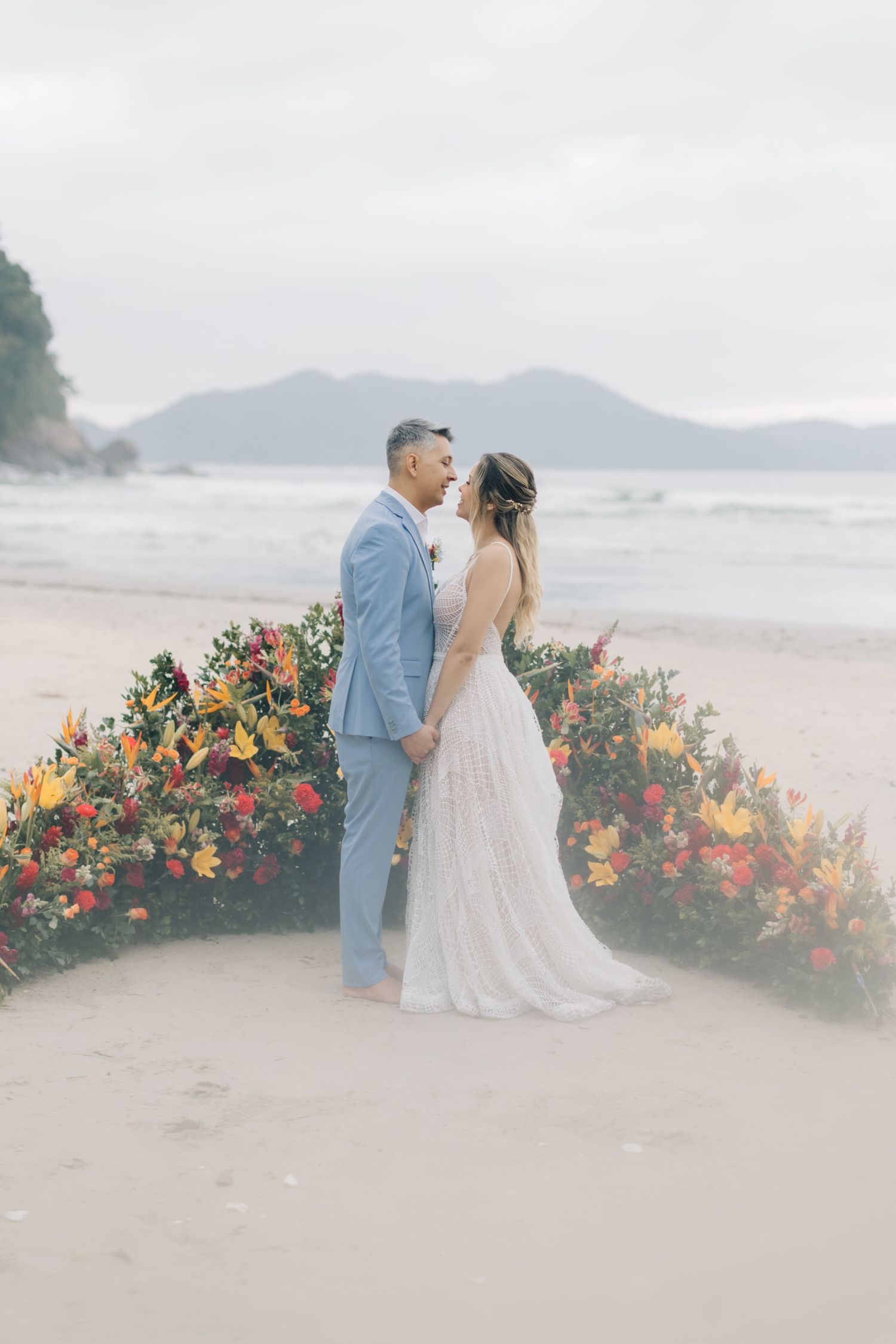 A couple shares an intimate moment during their beach wedding against a backdrop of colorful flowers and mountains.