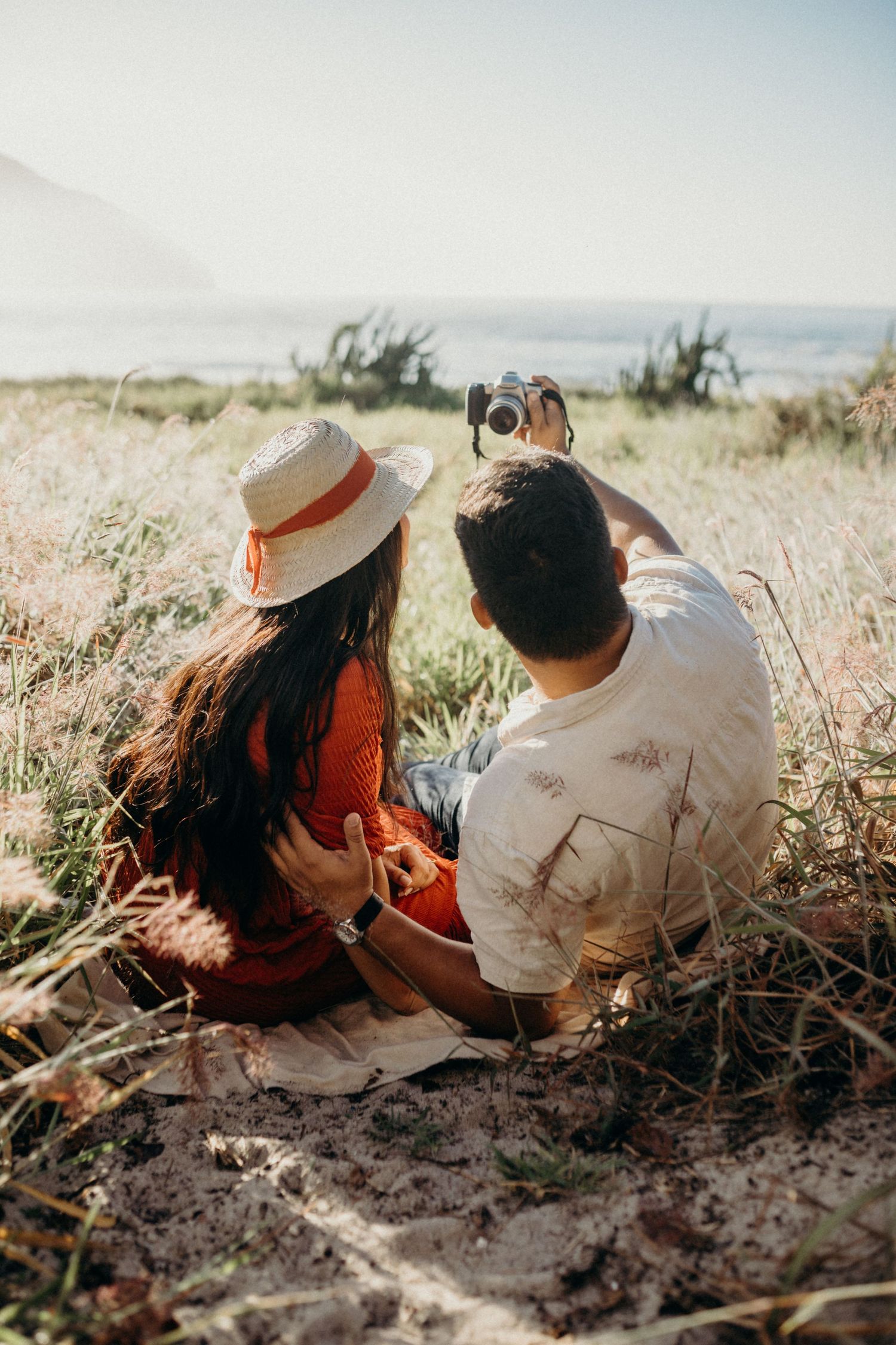 Two people sit close together on coastal cliffs with cameras capturing the ocean view.