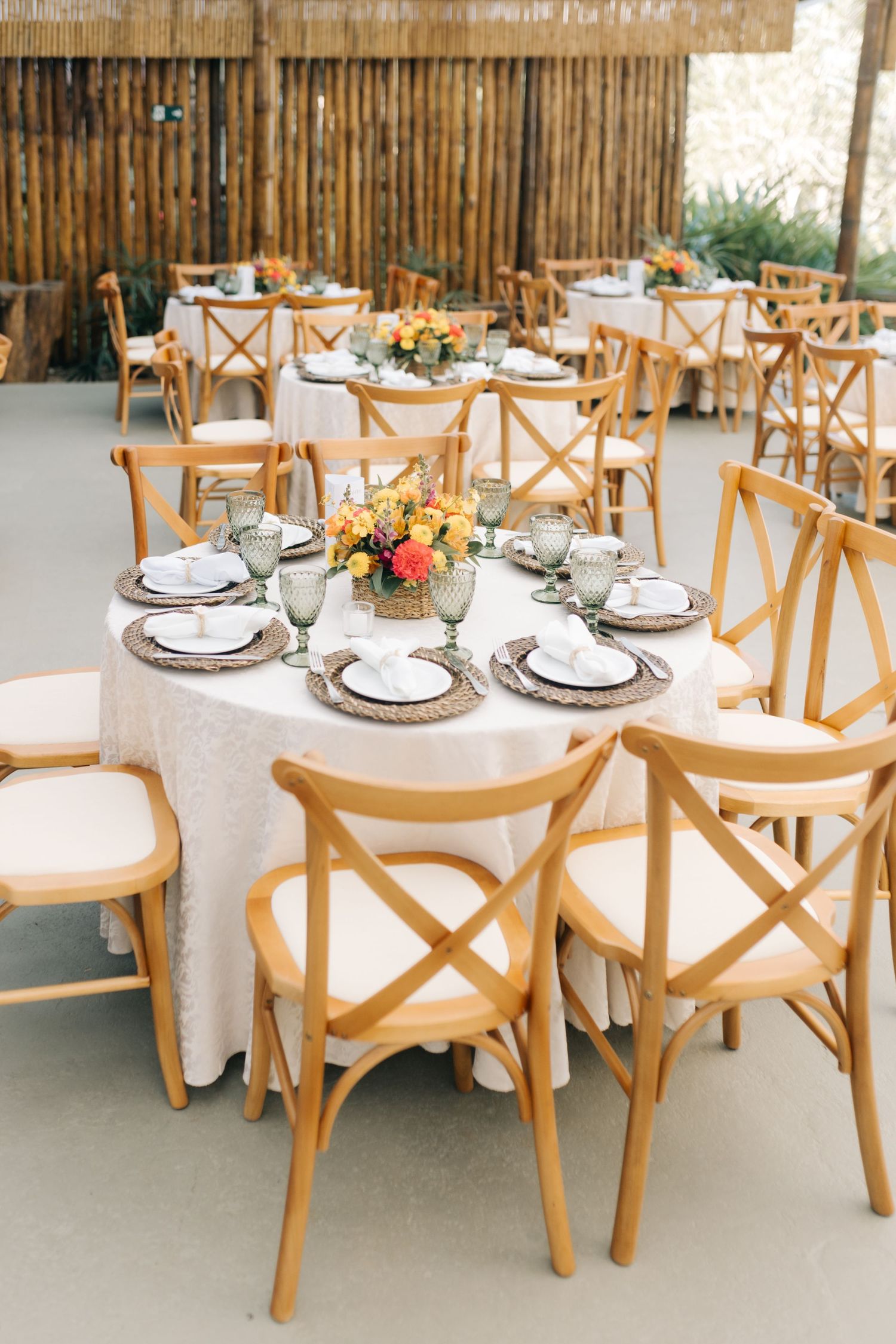 Wedding reception dining tables with wooden chairs and white tablecloths in a bamboo-walled venue.
