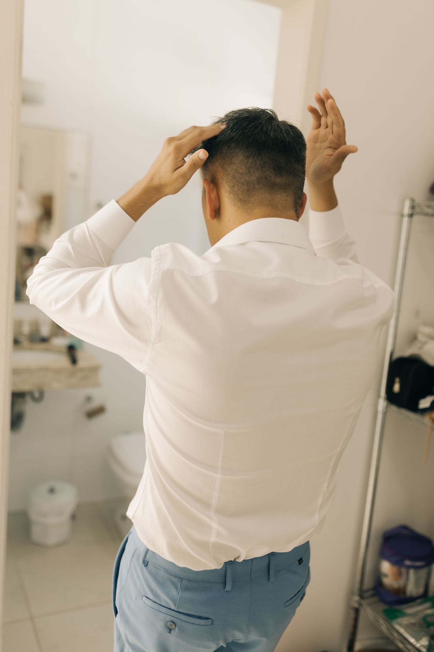 Man in white dress shirt and blue pants adjusts his hair while getting ready in a bathroom.