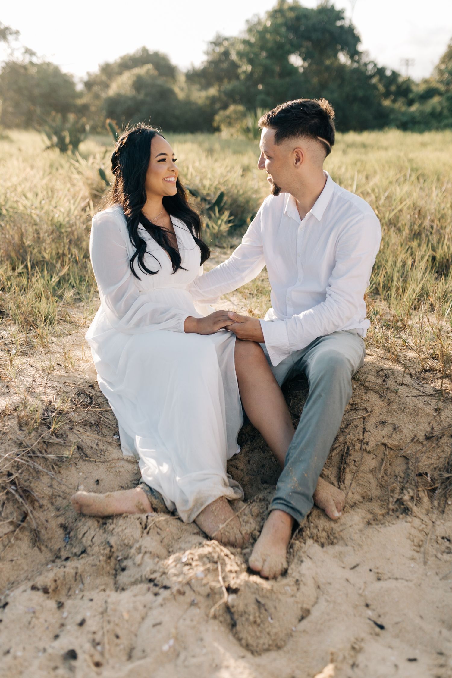 Romantic couple in white outfits share intimate moments while sitting on sandy dunes during golden hour.