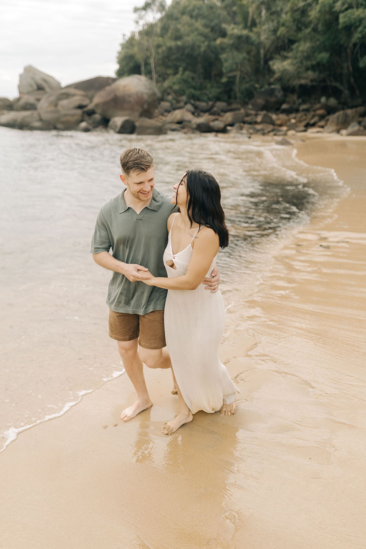 A couple stands together at the water's edge on a peaceful beach surrounded by rocky cliffs.