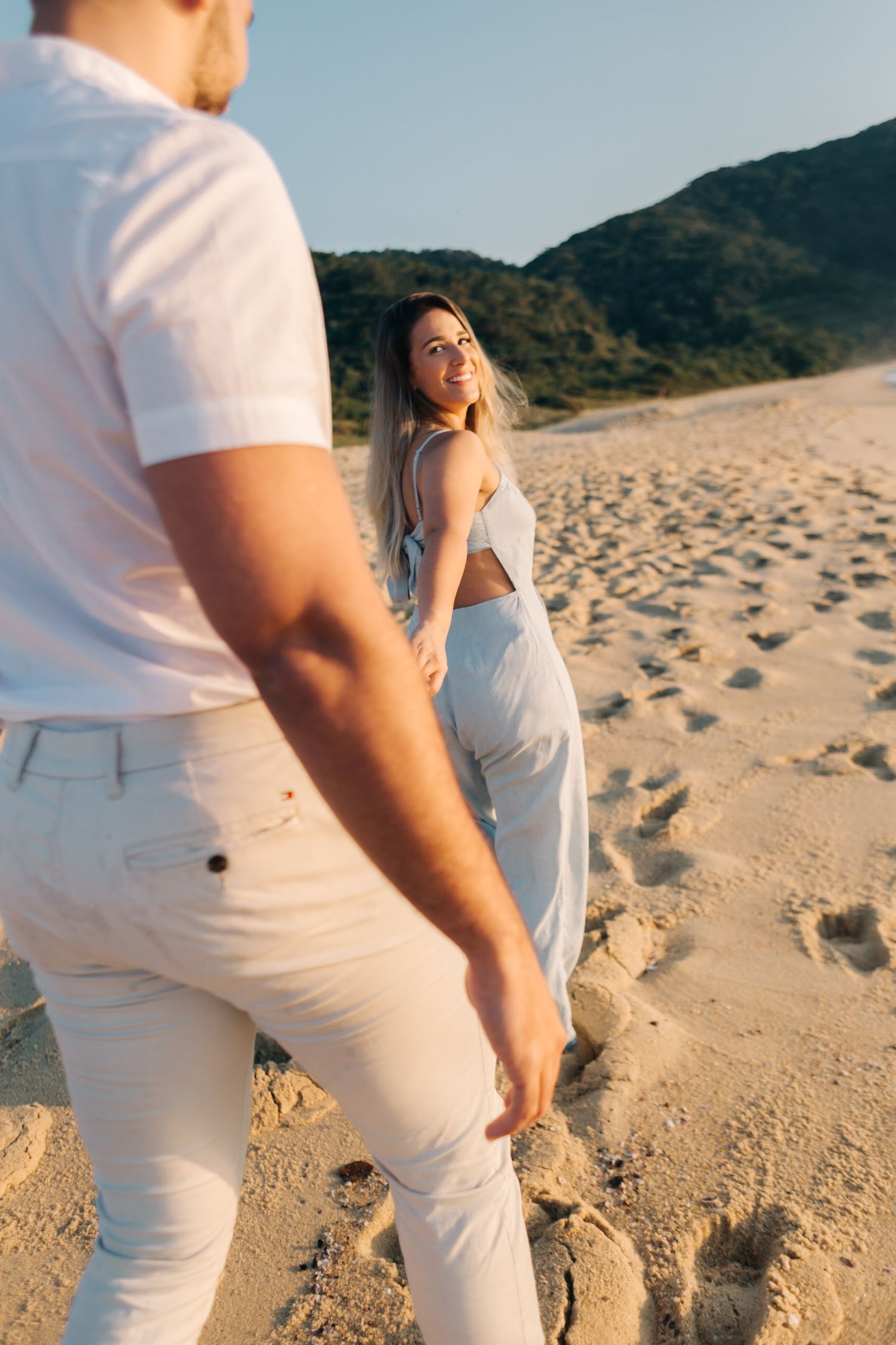 Couple in white outfits walking on sandy beach with mountains in background.
