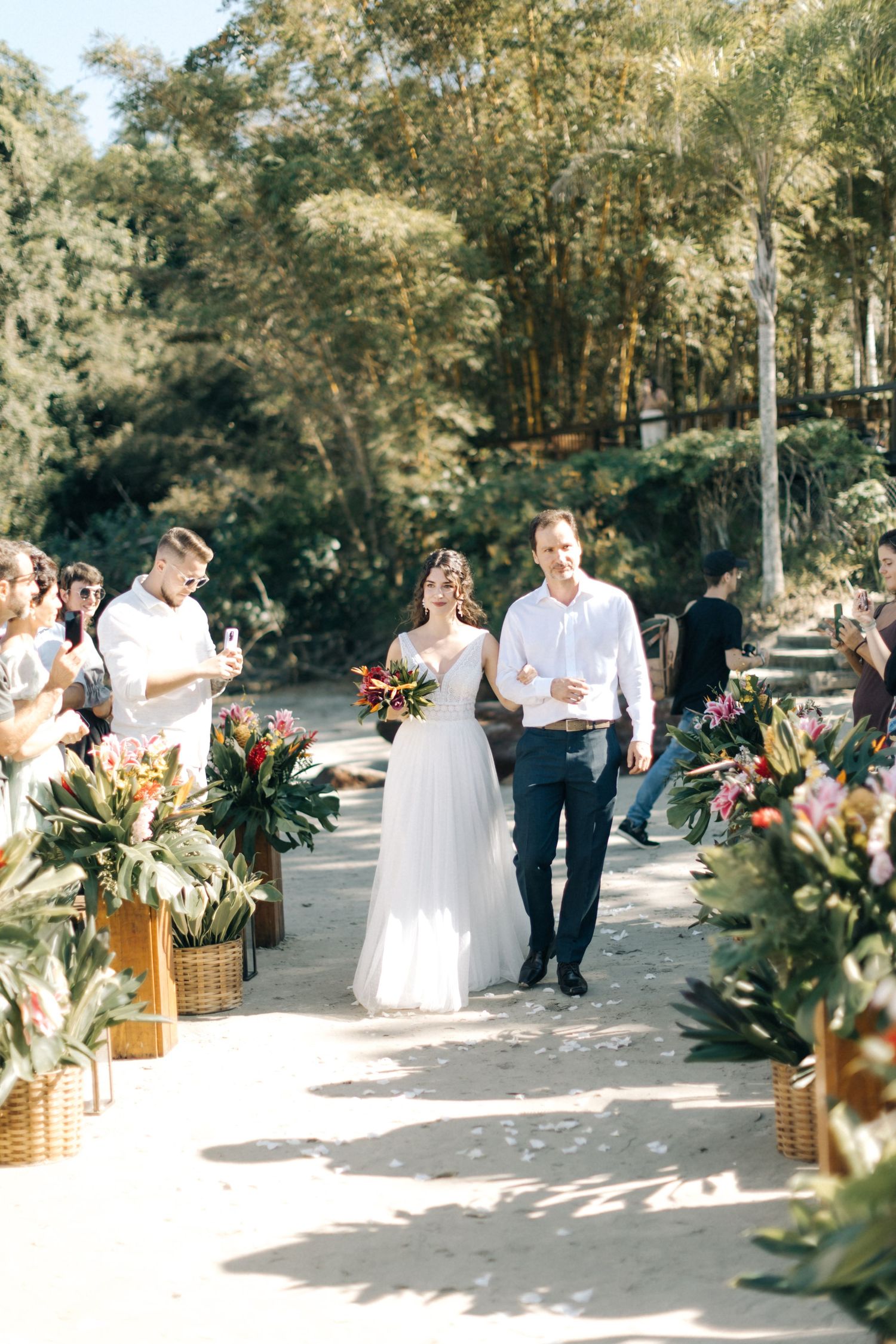 Wedding ceremony processional along a path lined with tropical plants and decorative boxes.