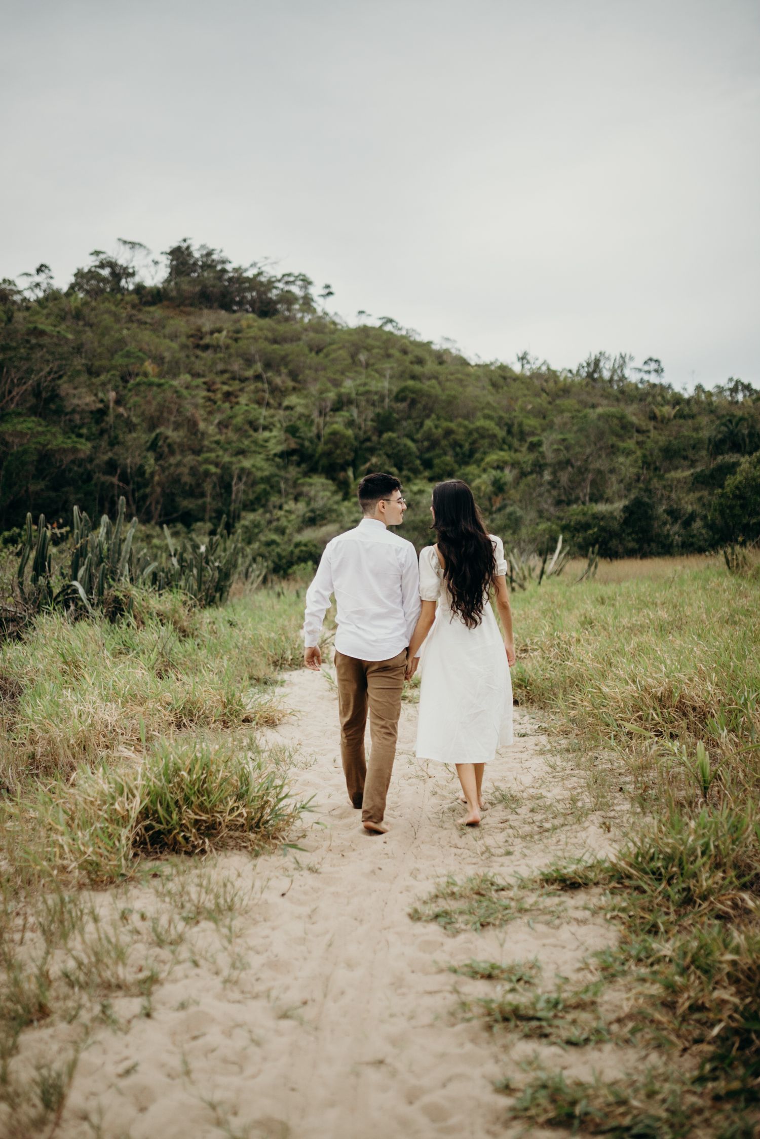 A couple walking hand in hand on a dirt path through grassy hills on a cloudy day.
