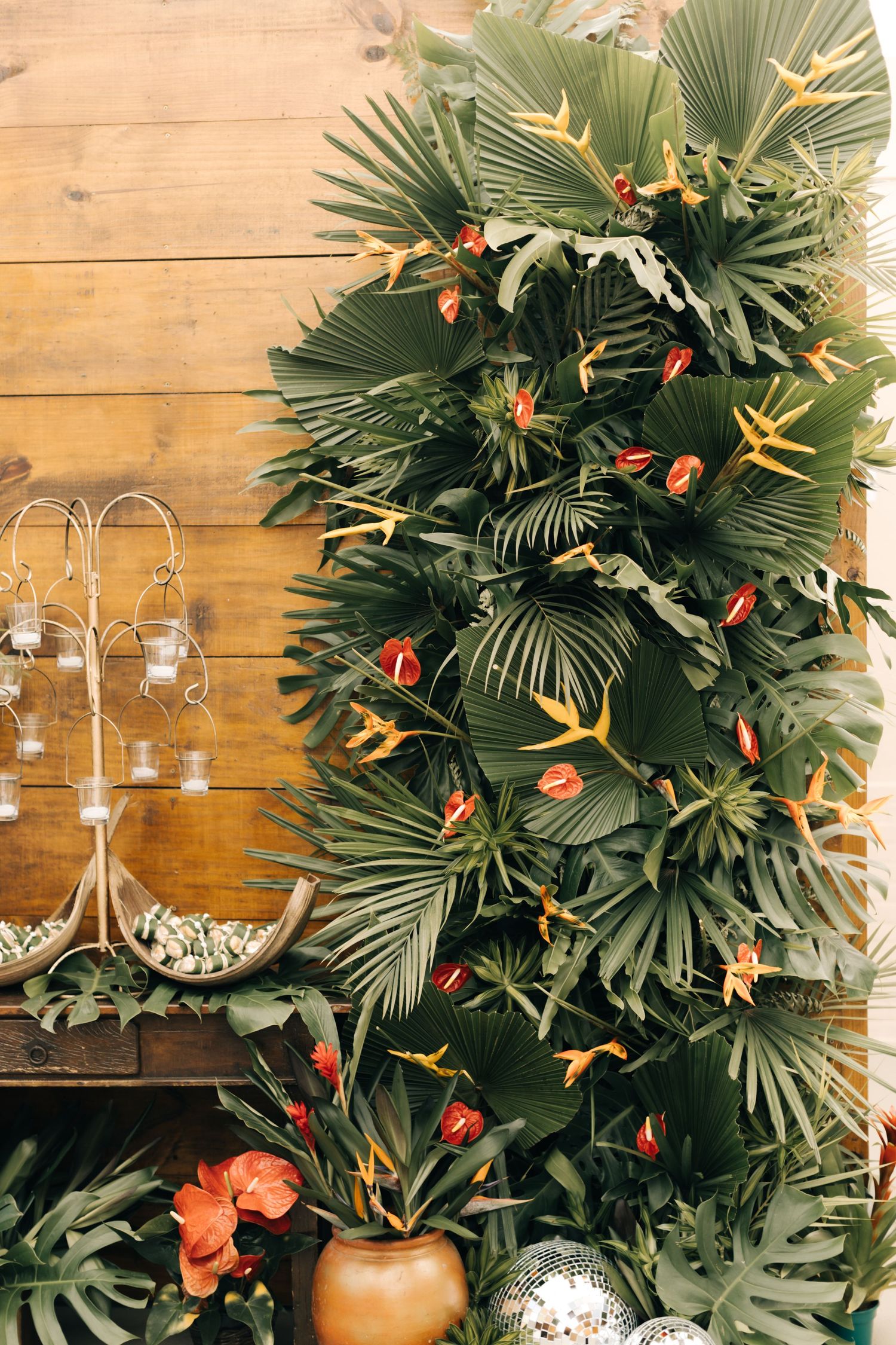 Lush tropical palm leaf wall installation with orange flowers and hanging glass candle holders.