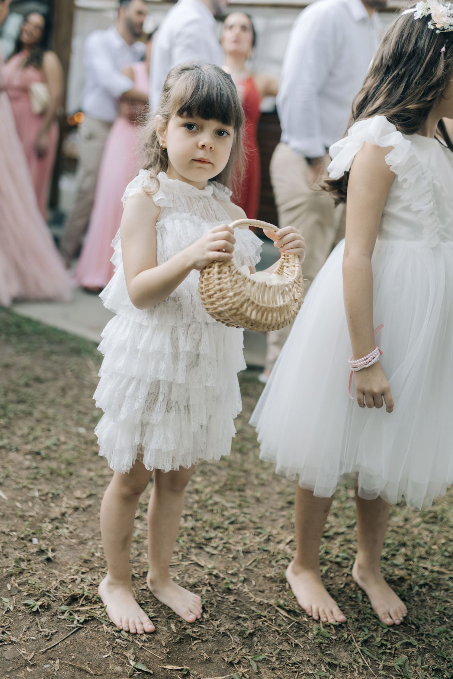 Young flower girls in white dresses hold wicker baskets at an outdoor wedding.