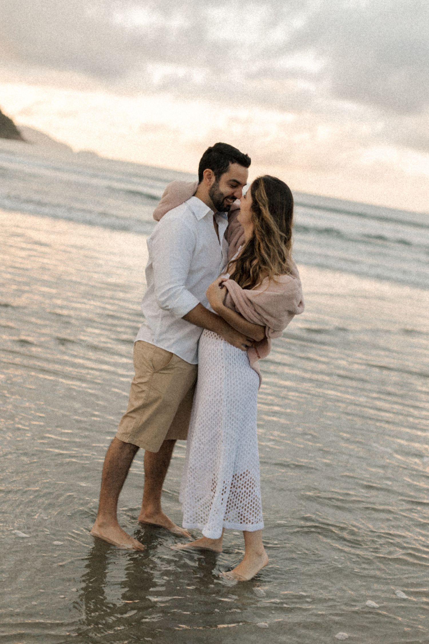 Romantic couple embraces on beach at sunset wearing white and beige casual attire.