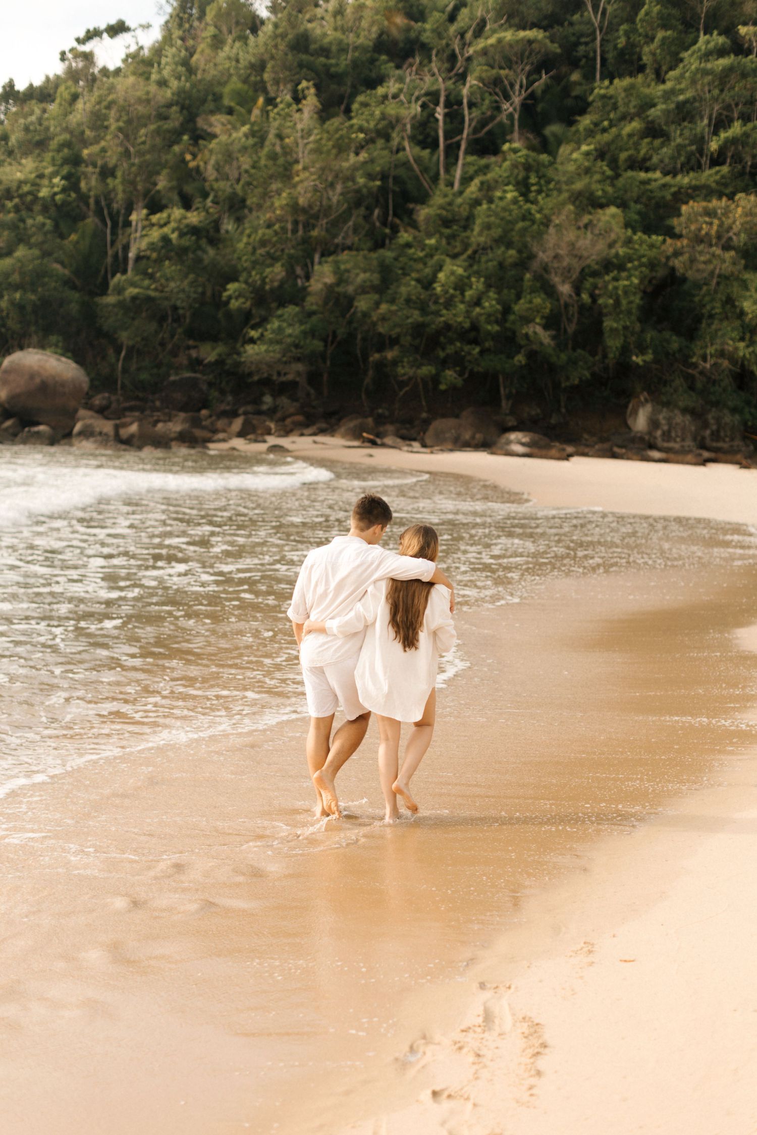 A couple wearing white clothing walk together intimately along a secluded tropical beach bordered by lush forest.