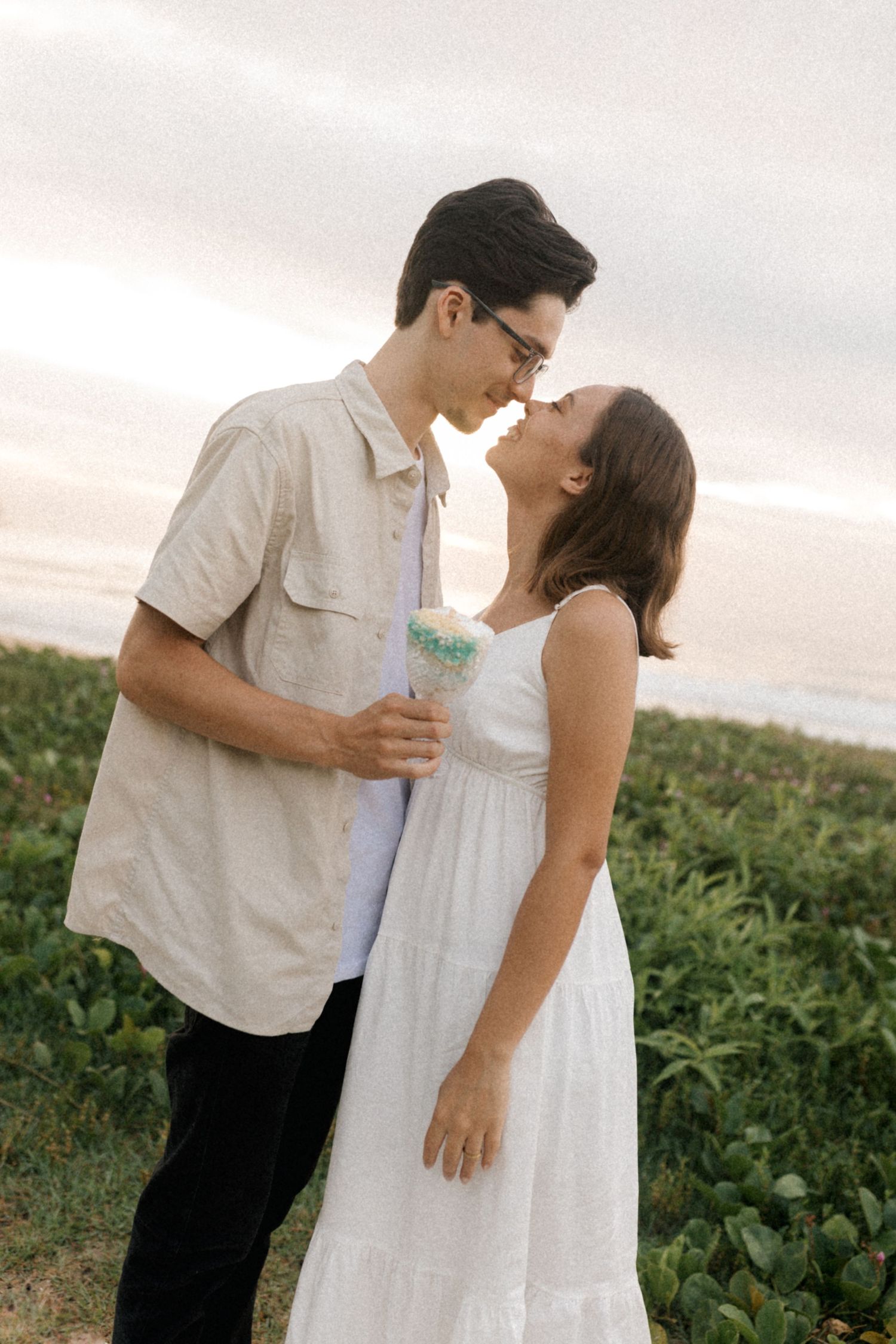 Sweet couple in white outfits shares tender moments and kisses while standing in a grassy field at dusk.
