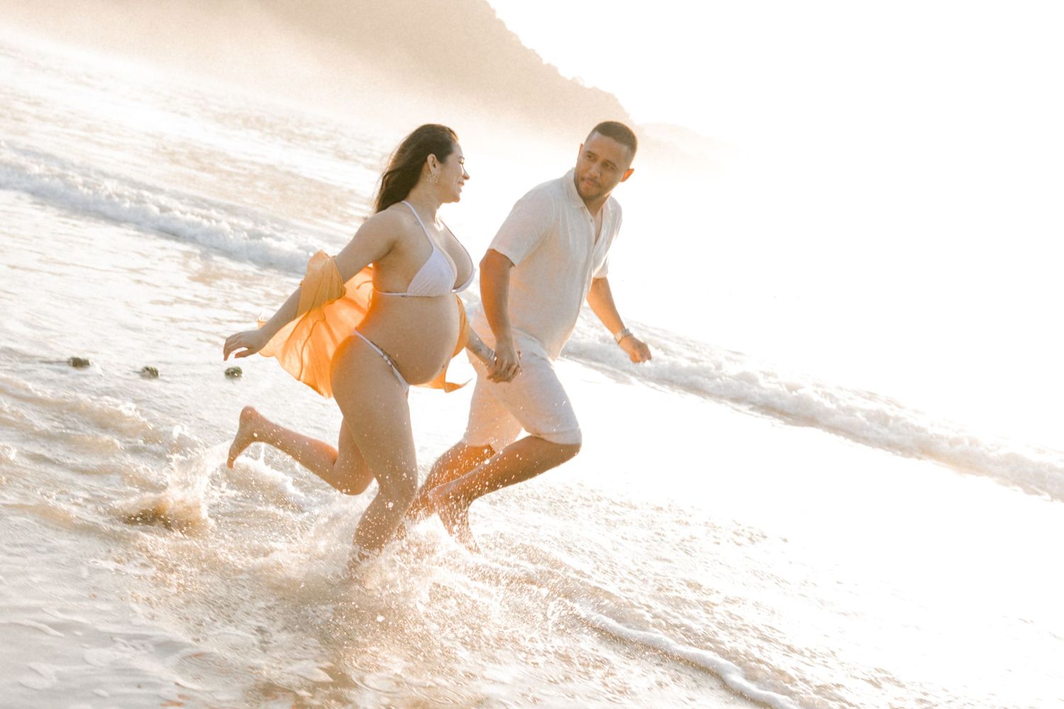 Couple runs through shallow ocean water during sunset wearing casual beach attire.