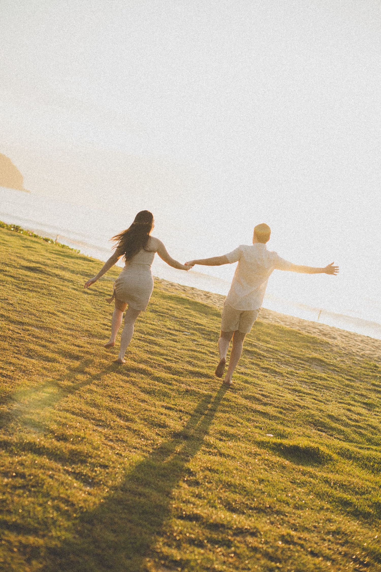 Couple running hand in hand on a grassy hillside during golden hour sunset.