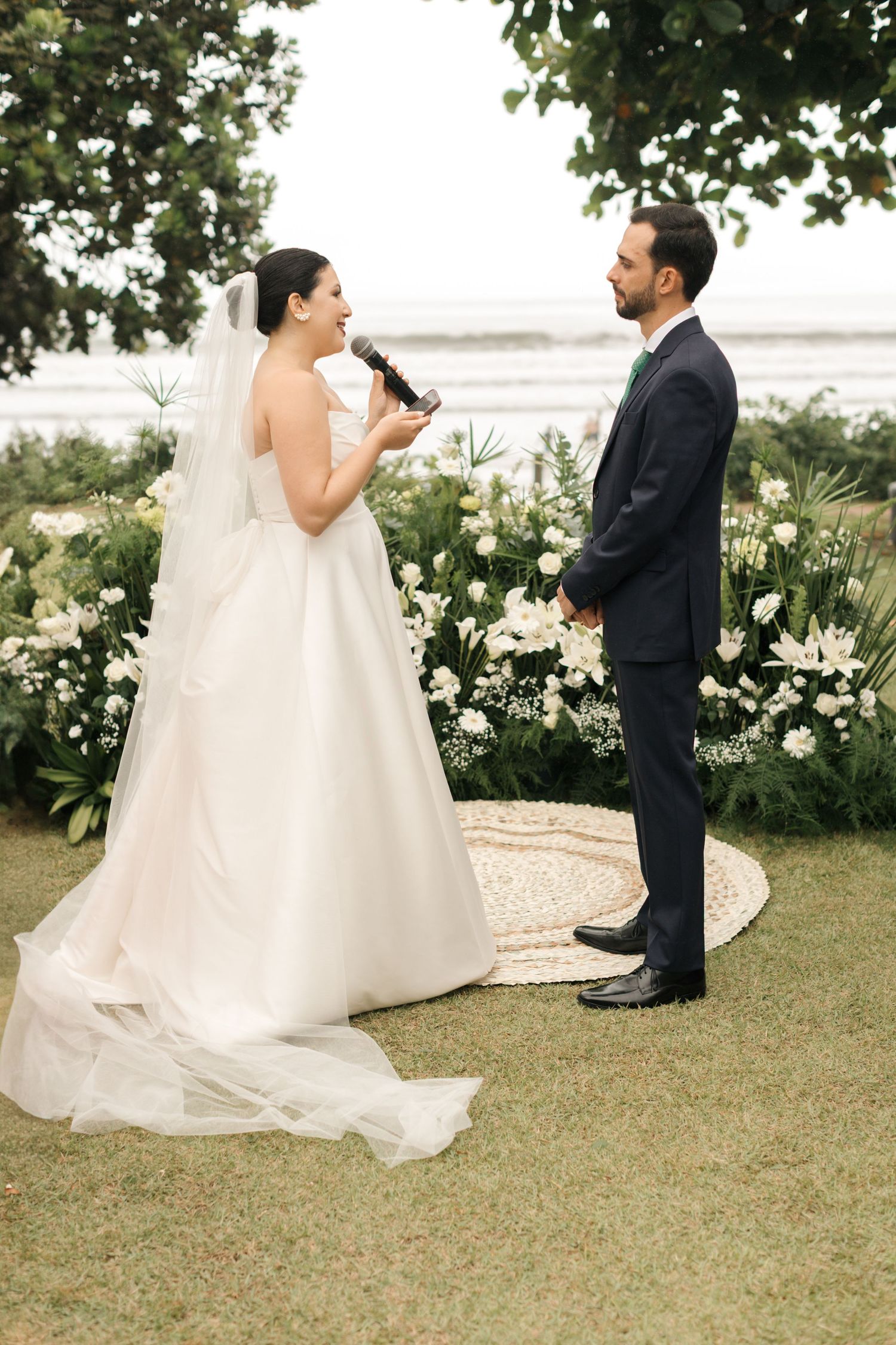 A wedding ceremony moment with the bride speaking into a microphone outdoors.