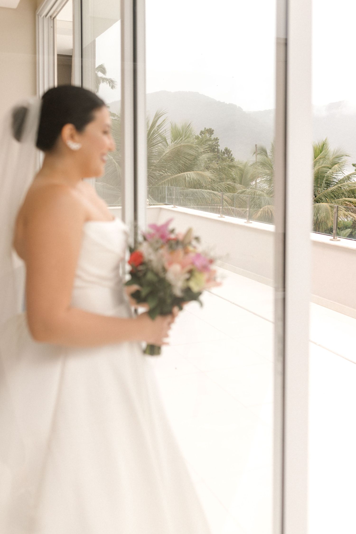 A bride in a strapless gown holds a bouquet while looking out a window with palm trees visible.