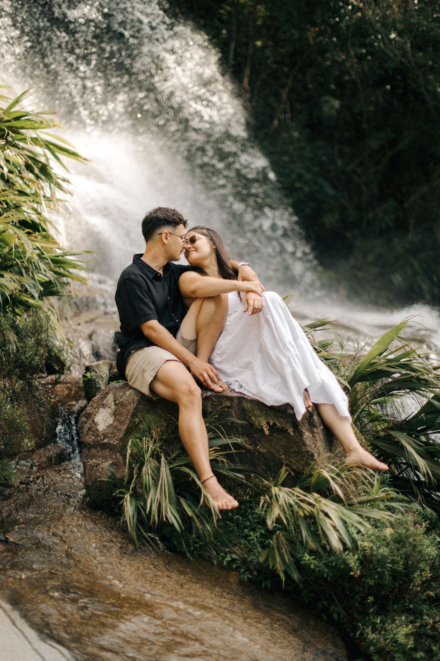 Tender moment photographed beside a majestic waterfall with natural greenery surrounding.