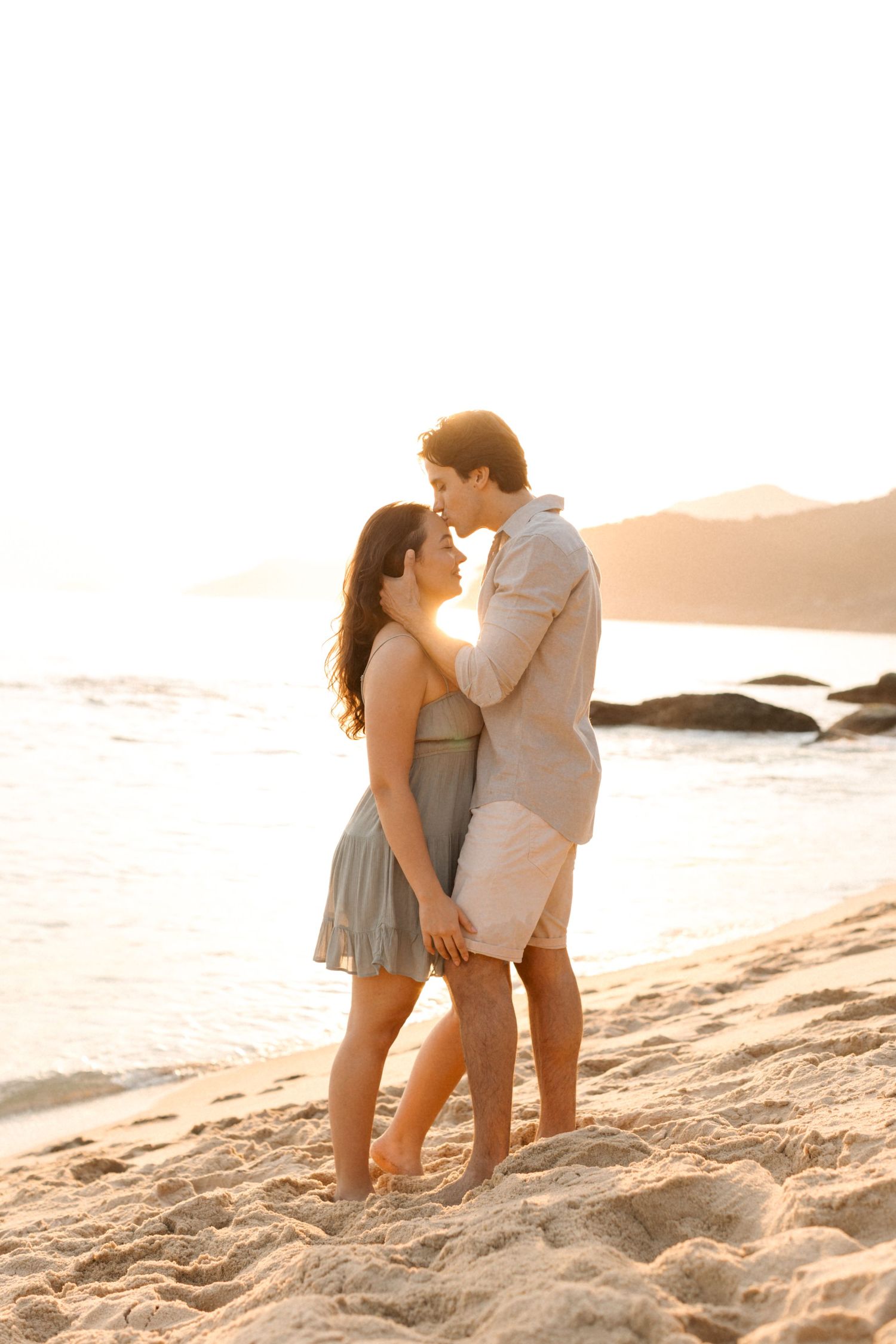 Couple embracing on sandy beach with ocean waves and sunset in background.