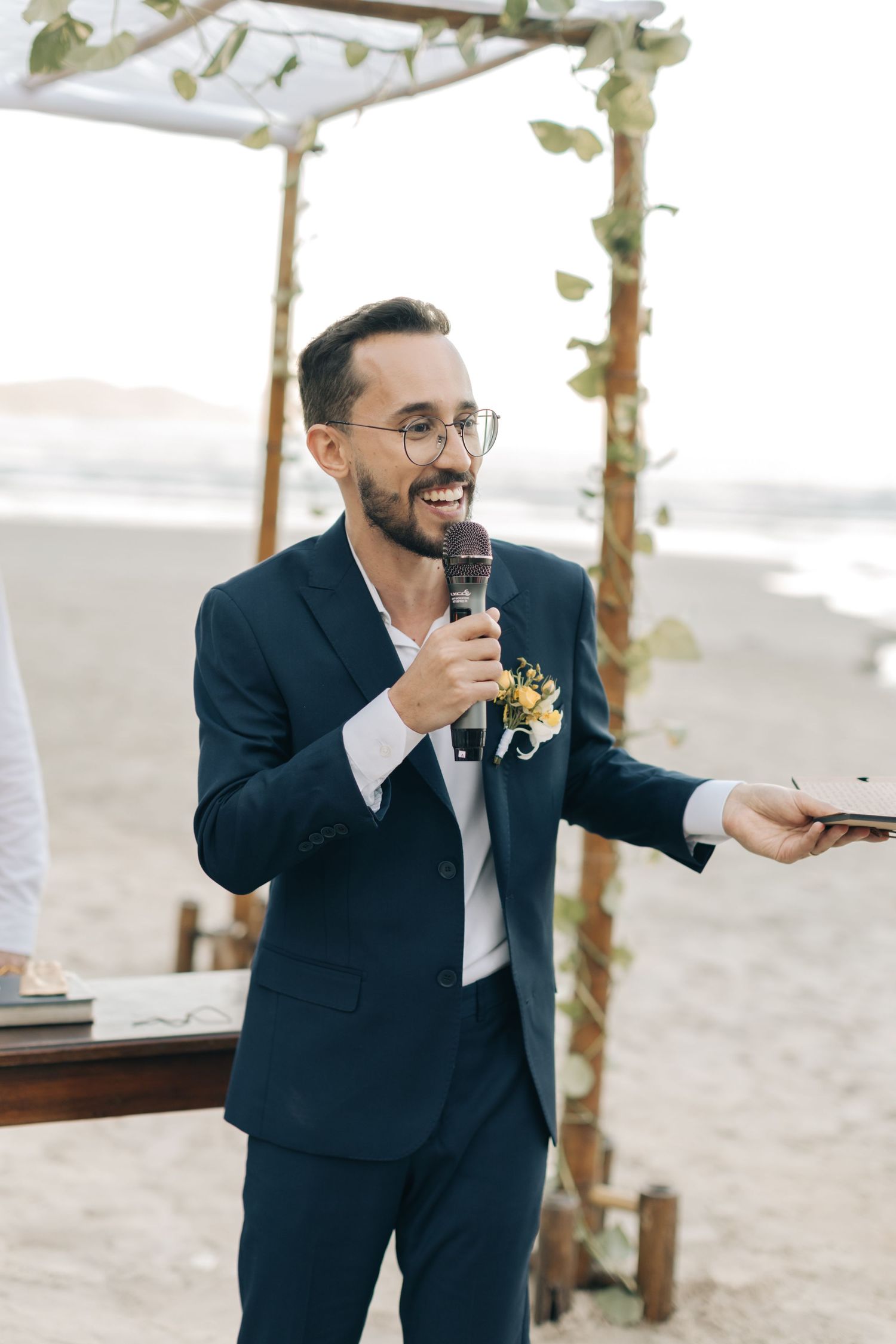 Groom in navy suit makes a speech while holding a microphone at an outdoor beach ceremony.