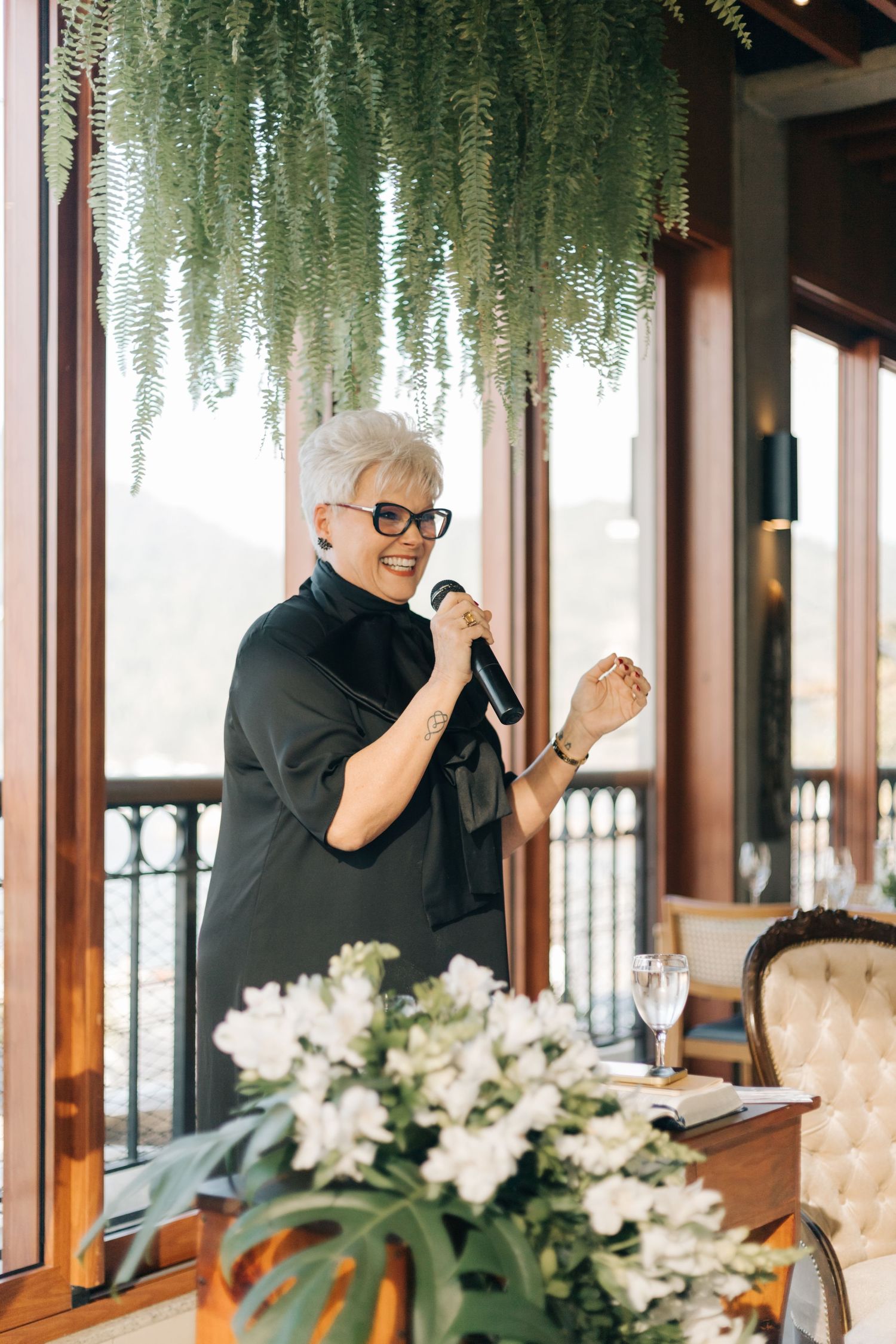 Multiple images of a speaker in black addressing guests at a wedding venue with hanging greenery.