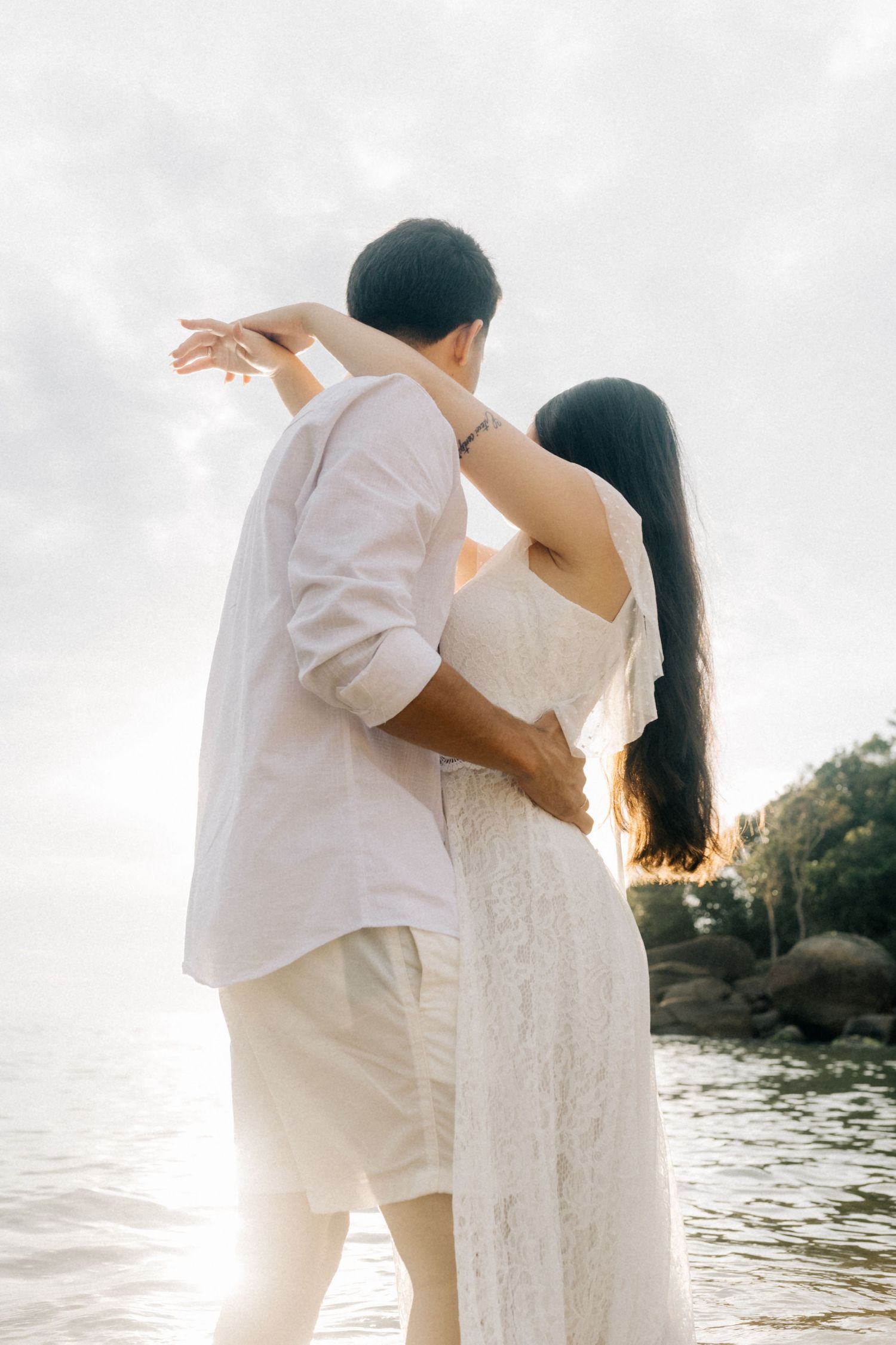 Elegant couple in white outfits pose together near water's edge.
