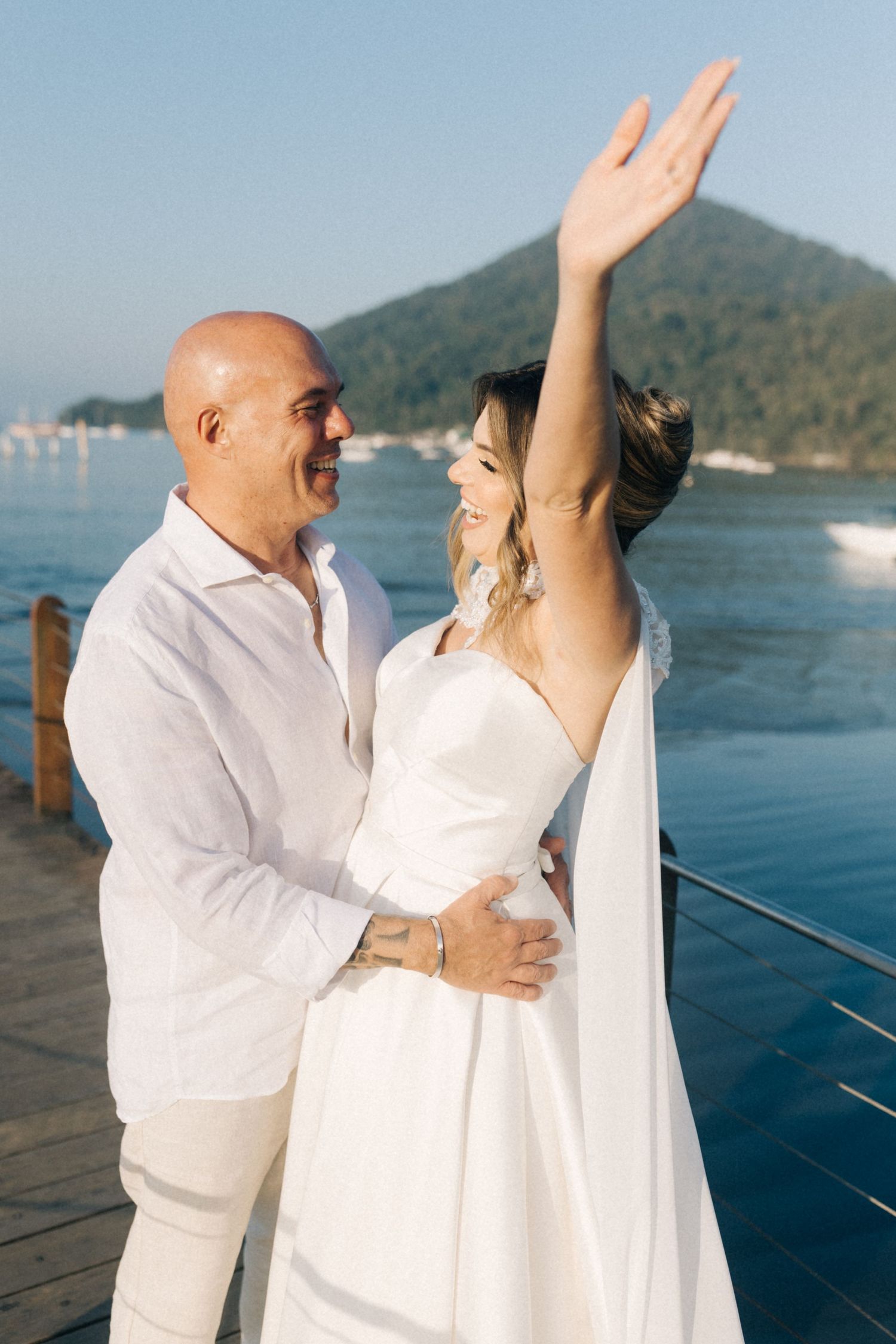 A couple shares a joyful moment on a dock with mountains and ocean in the background.