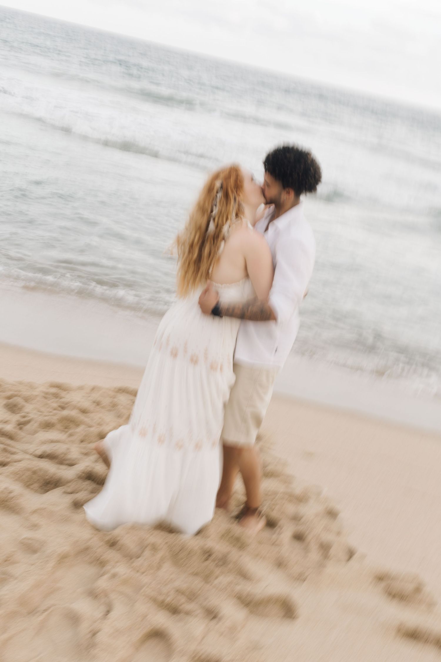 A couple in light colored outfits embrace while standing on a beach at sunset.