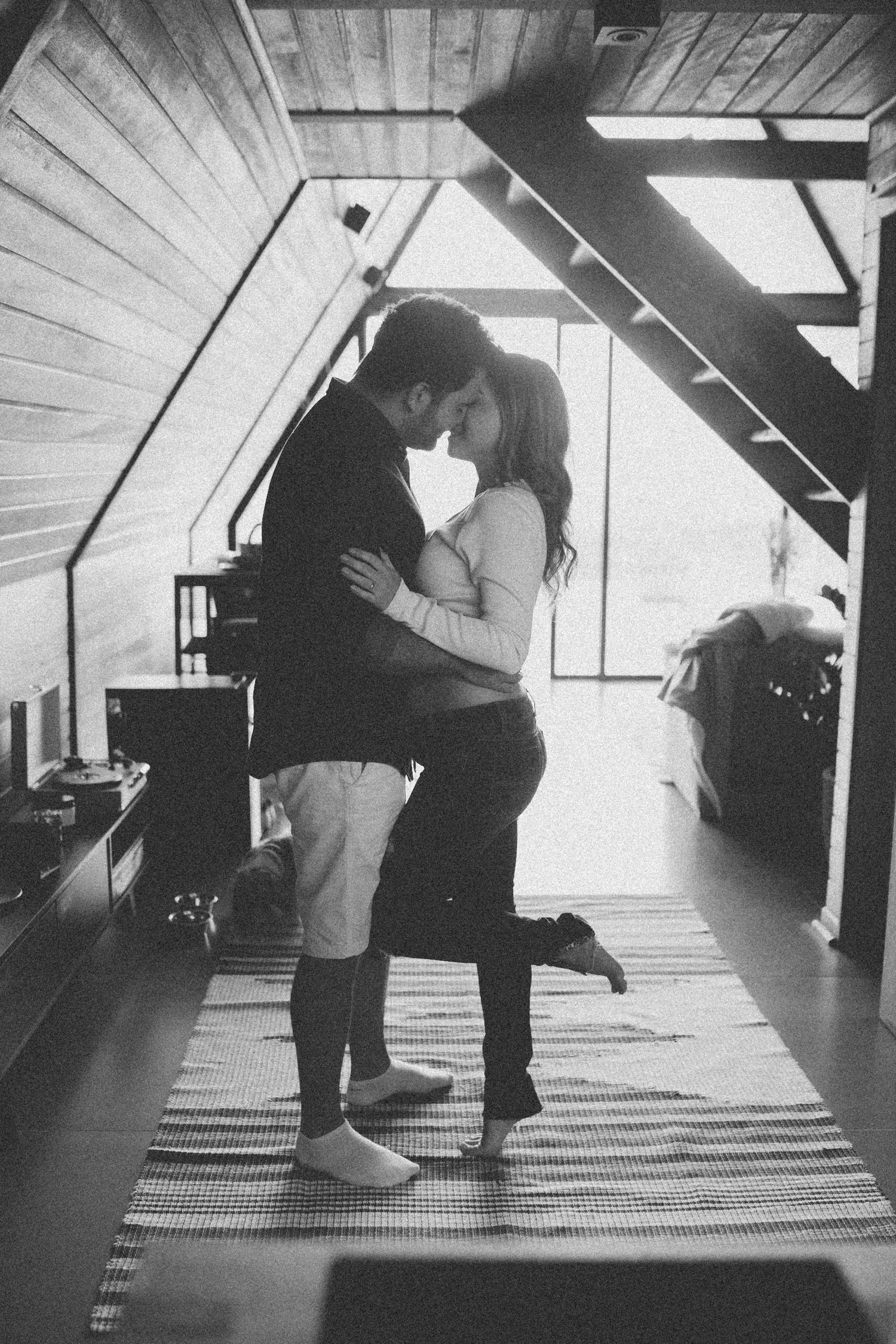 A romantic embrace captured in black and white within a sunlit wooden attic space.