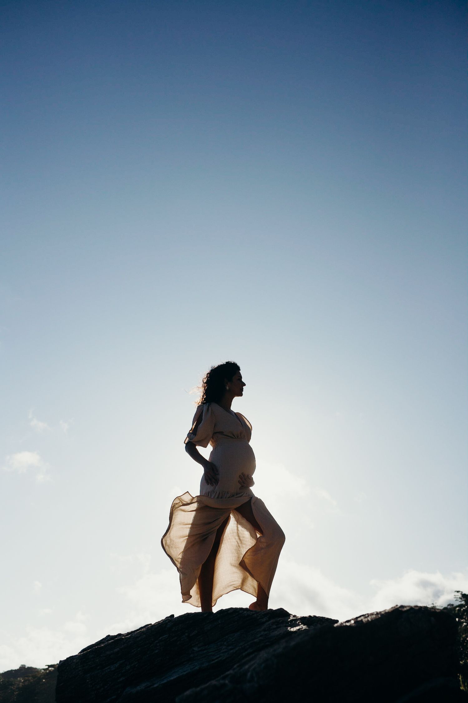 Expectant figure poses on rocky overlook against bright sky.