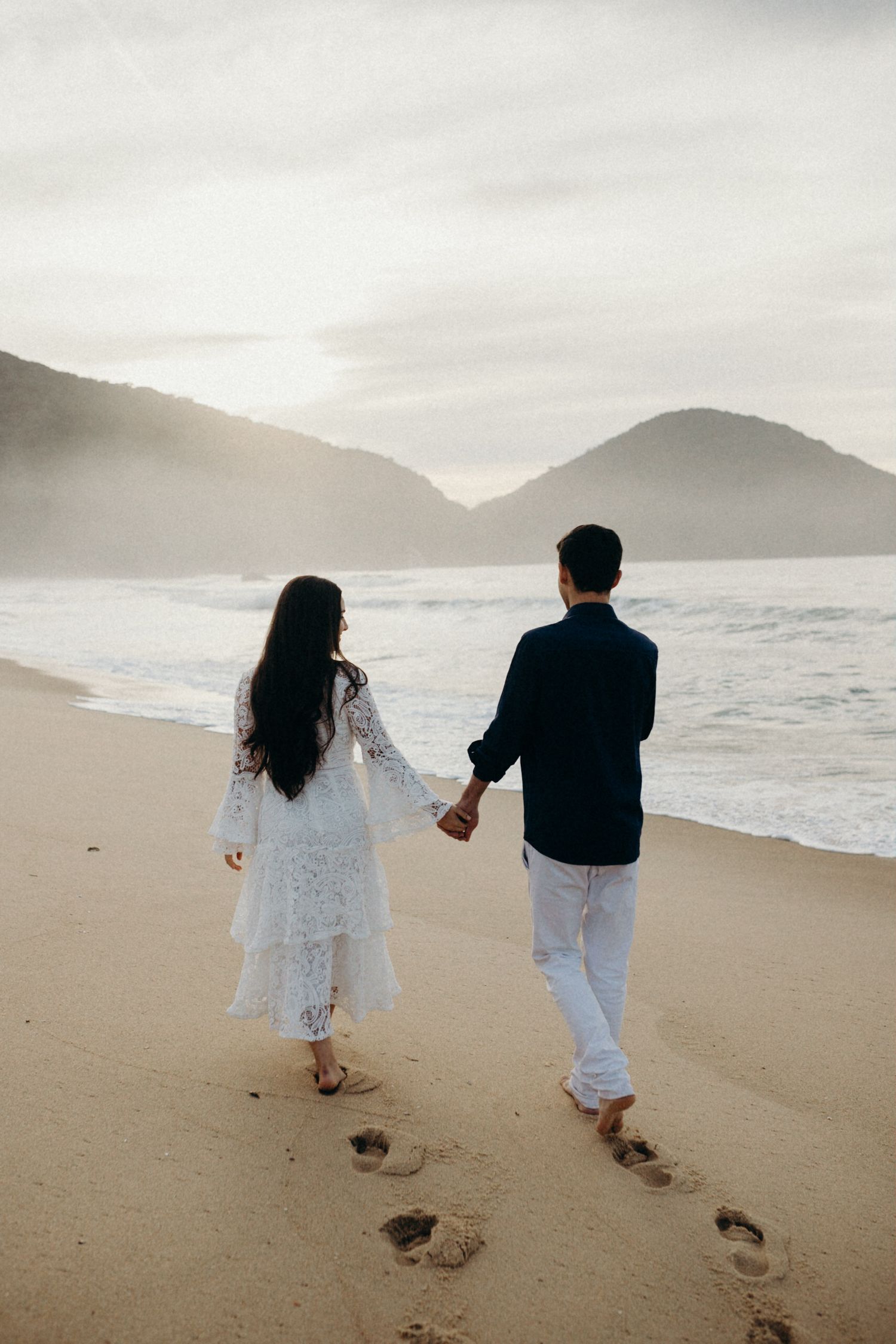 Couple walking hand in hand on beach leaving footprints in sand with mountains in background.