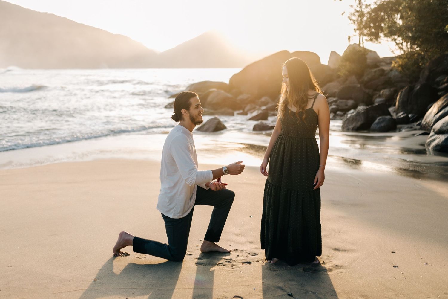 A couple sharing an intimate proposal moment on a secluded beach with rocky shoreline.