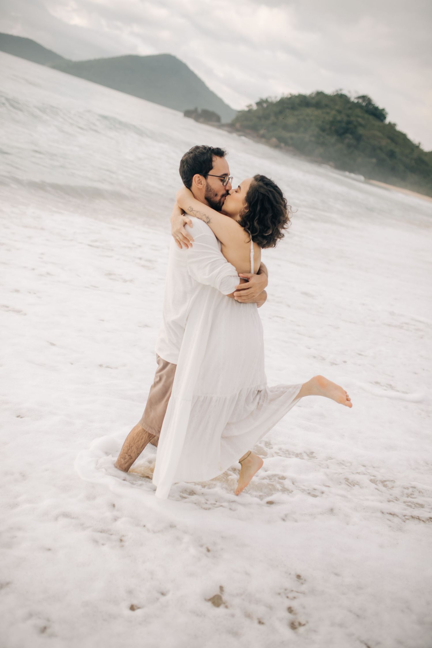 A romantic couple embracing on a misty beach wearing white outfits by the ocean.