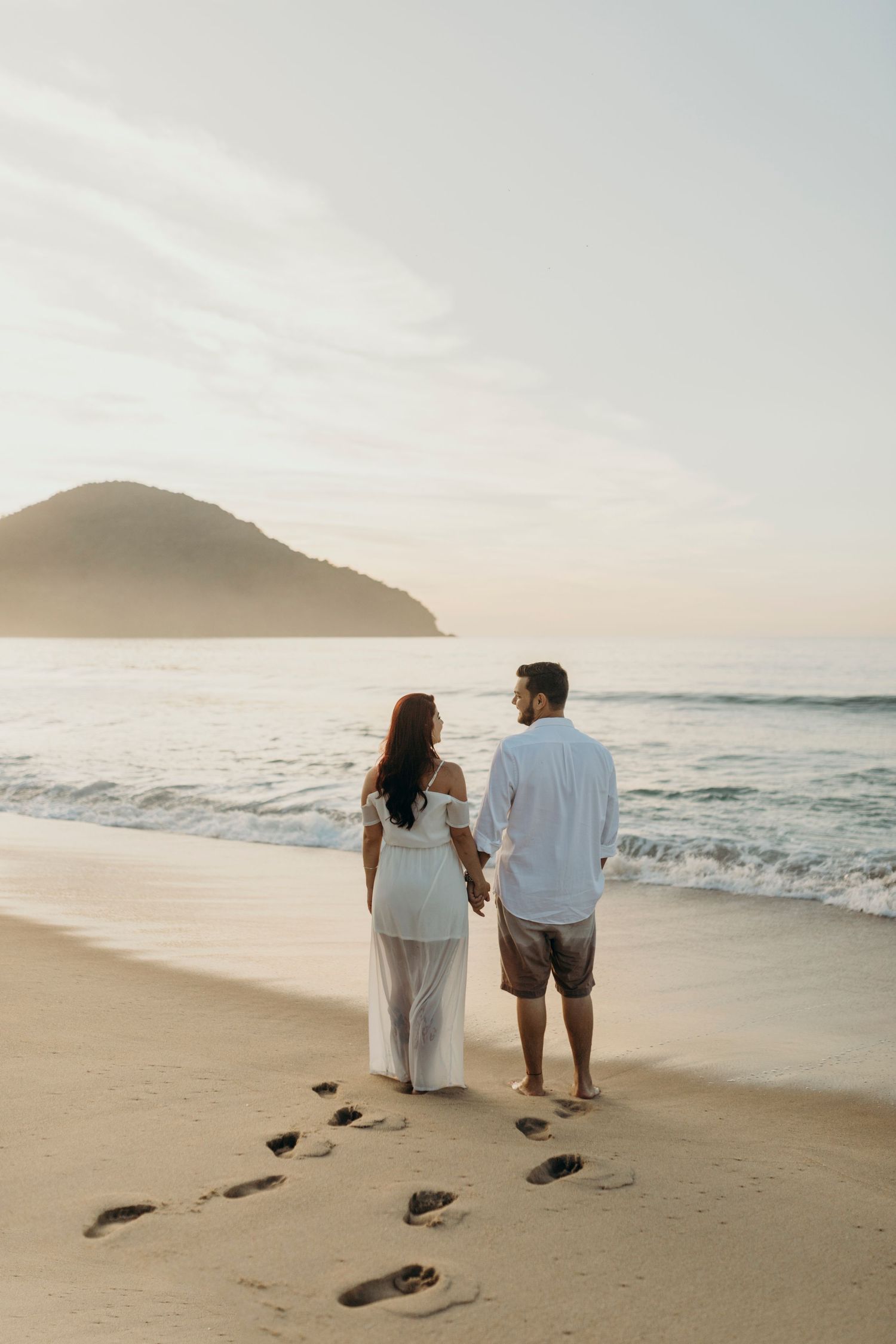 A couple leaves footprints in the sand while walking along a beach at sunset.