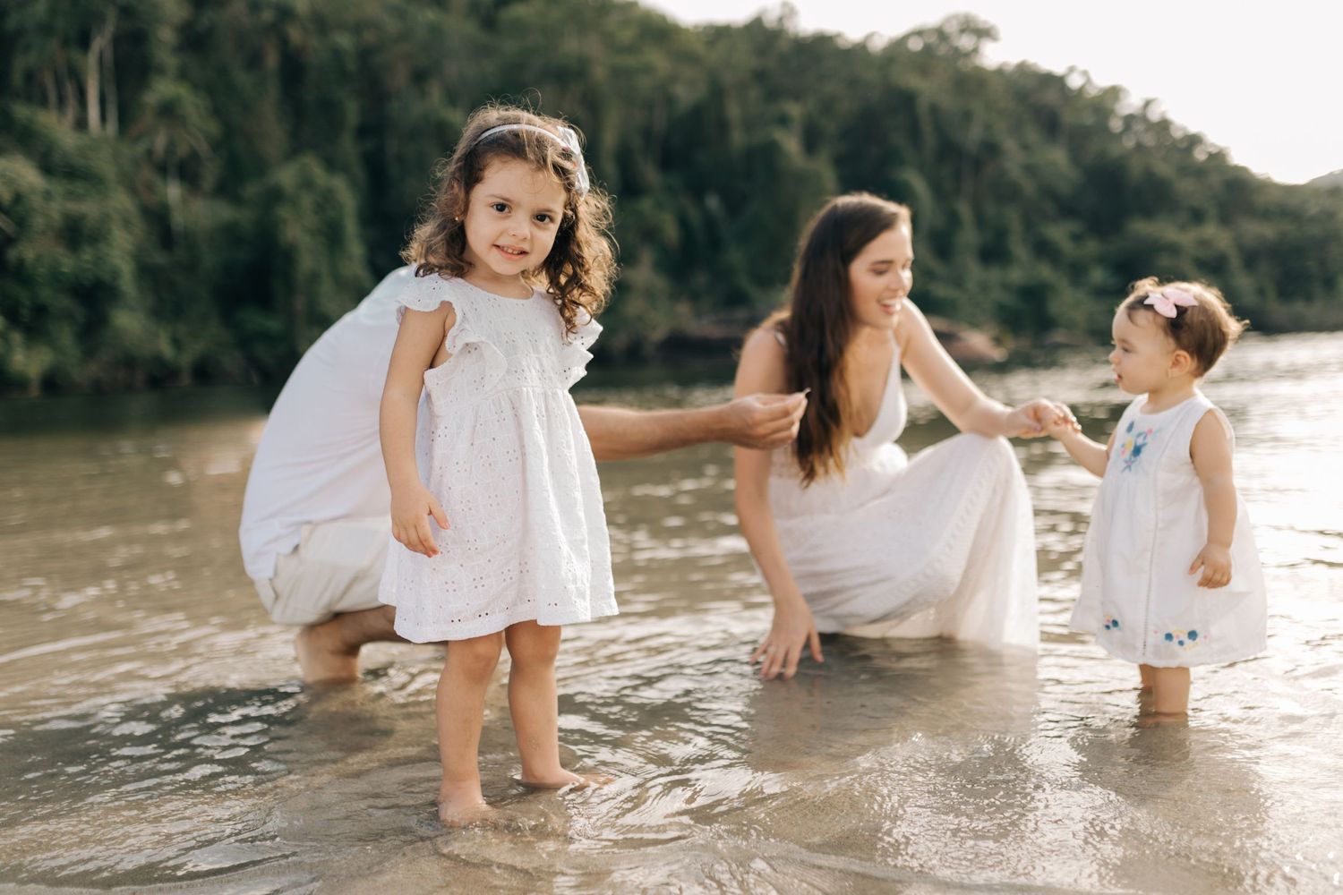 Family enjoying a playful moment together at water's edge in white summer clothing.