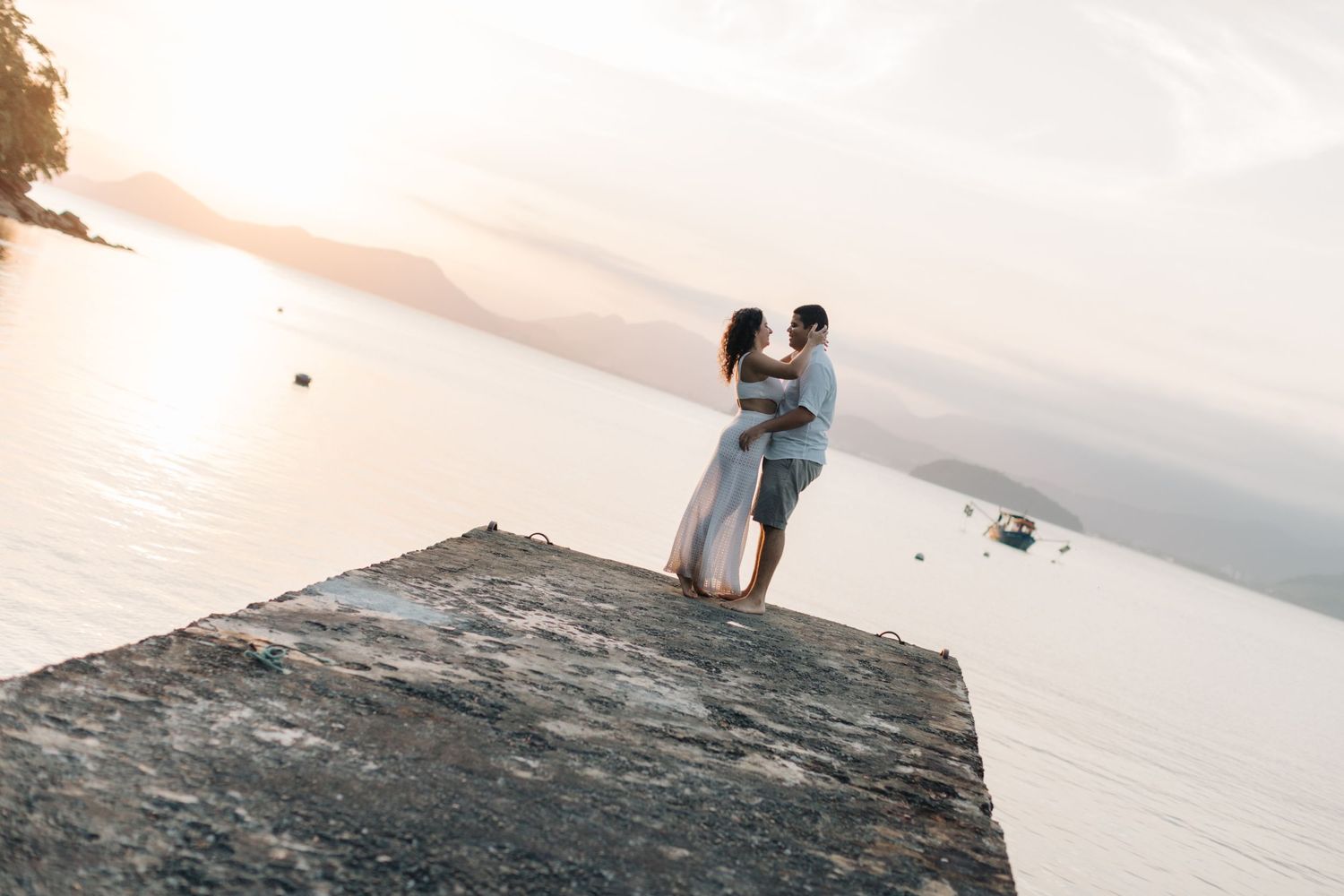 Aerial view of a couple embracing on a concrete pier overlooking the ocean at sunset.