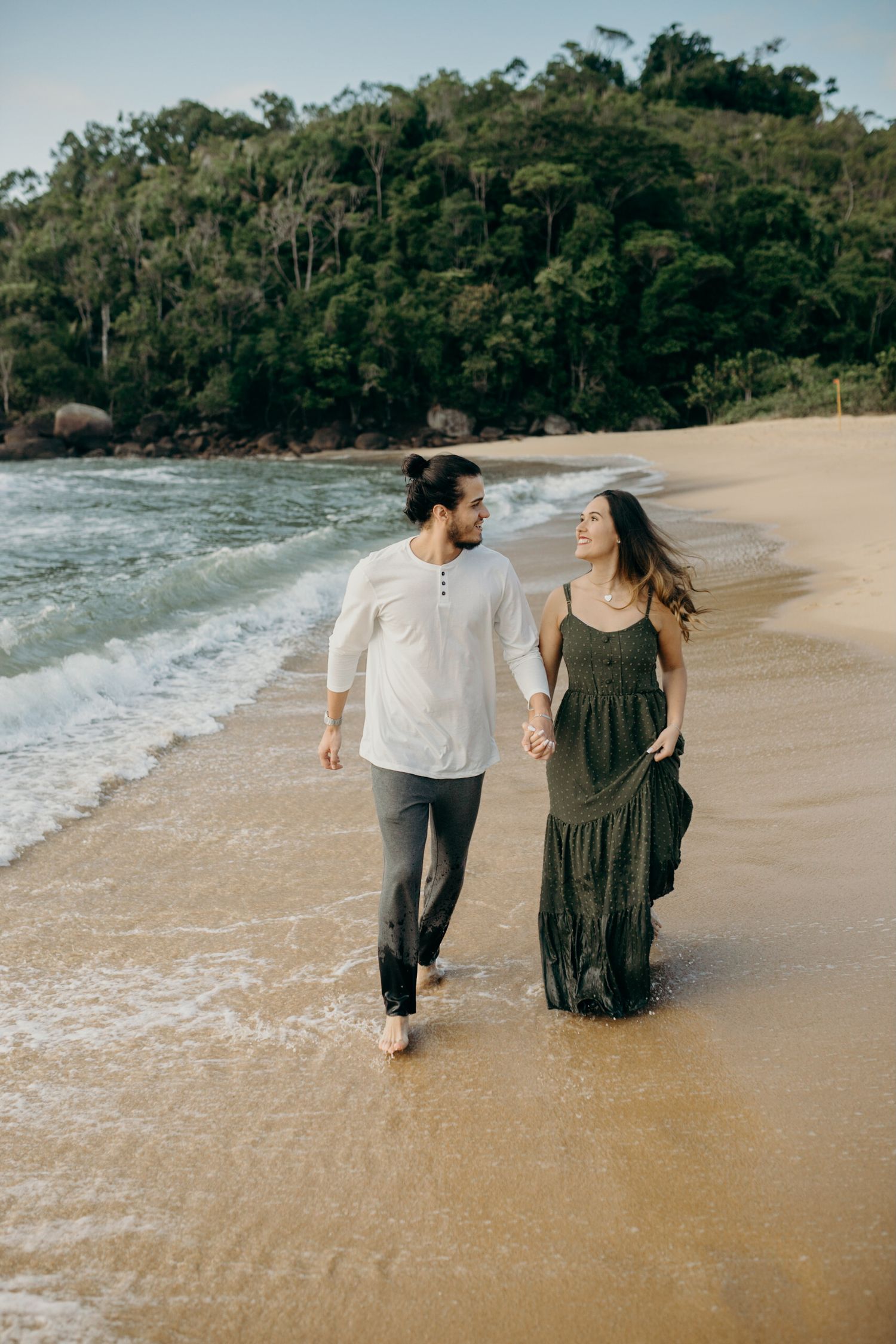 Couple strolls together on a sandy beach with waves and forested coastline in the background.